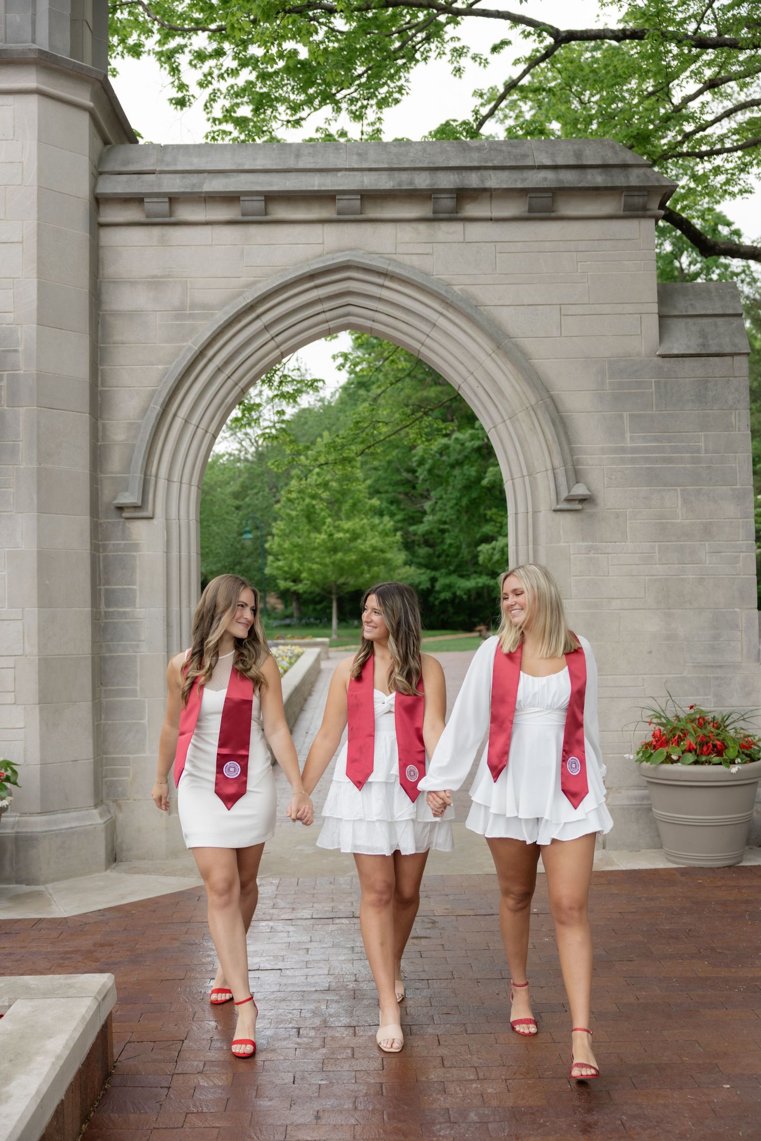 Three women in white dresses and red accessories walk together along a stone archway path.