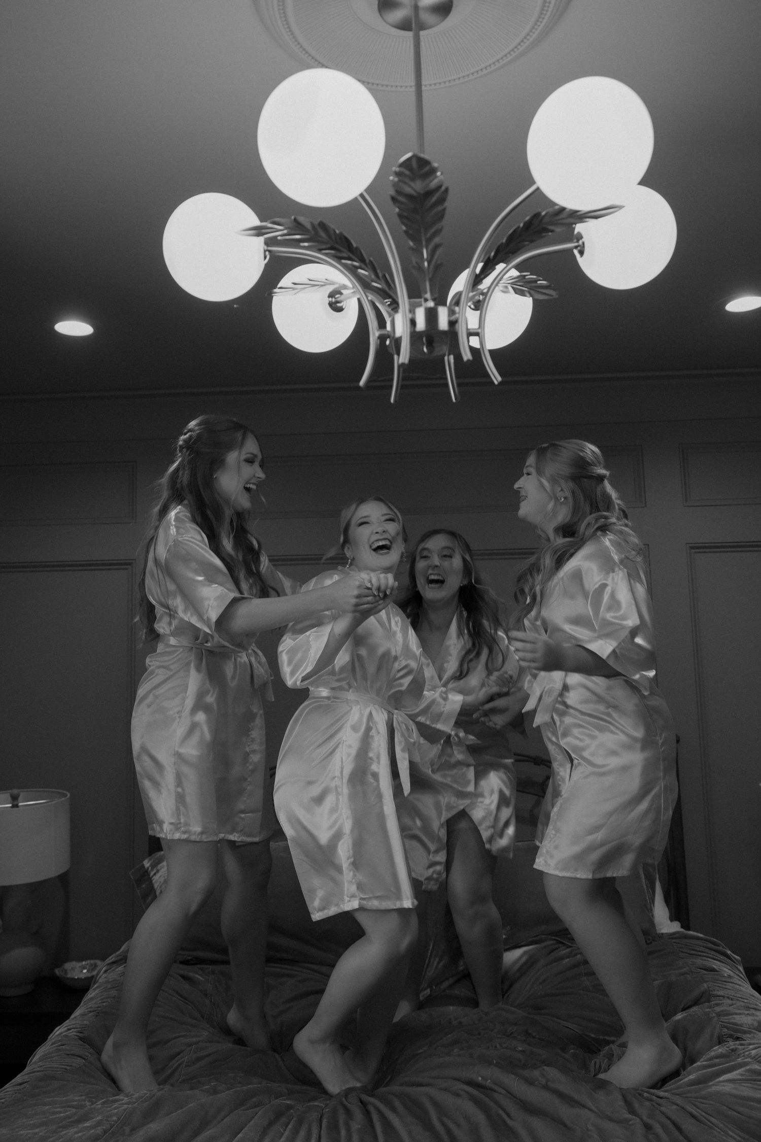 Group in white satin robes having fun jumping on a bed in a room with round lights.