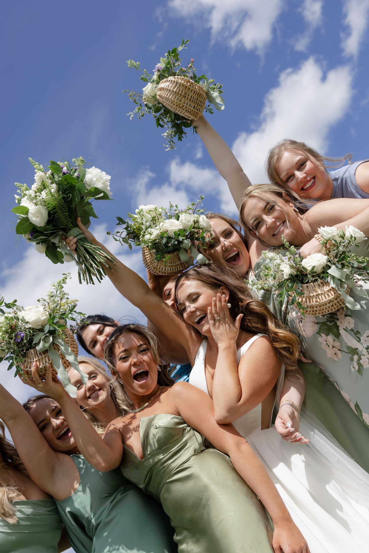Wedding party in sage green dresses celebrating joyfully against a bright blue sky.