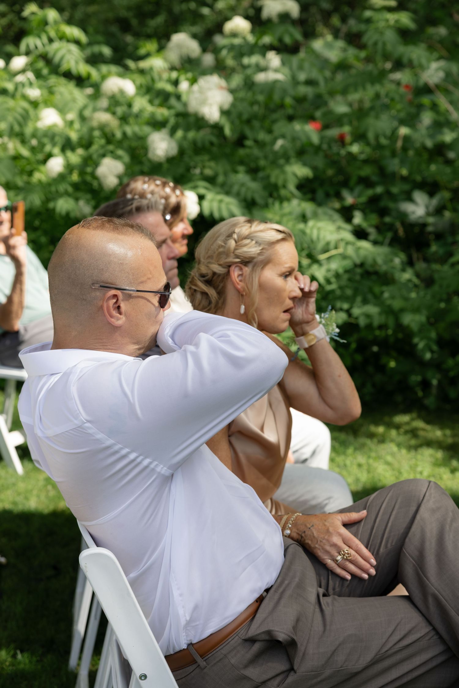 Guests in white and grey attire seated outdoors watching an emotional ceremony moment.
