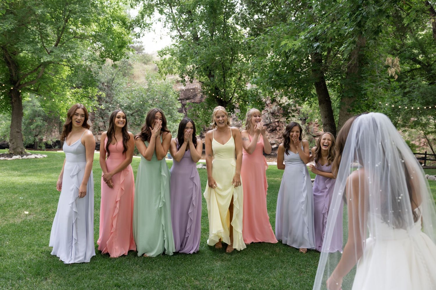 Bridal party in pastel dresses poses together outdoors under trees.