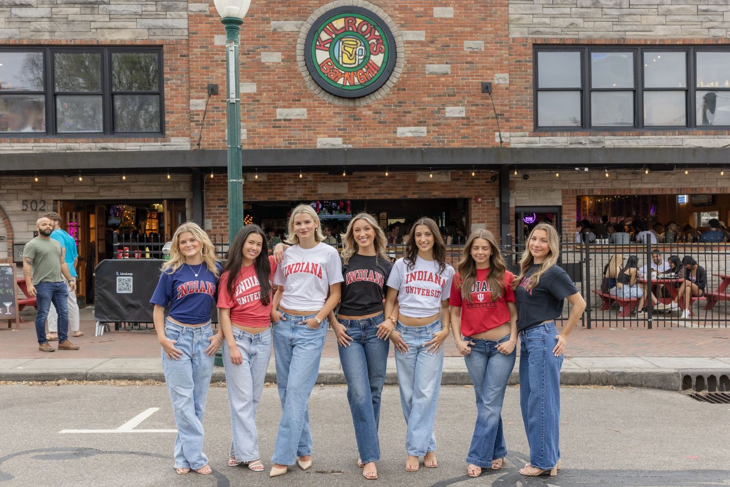 Group in matching collegiate t-shirts and jeans pose outside campus building.