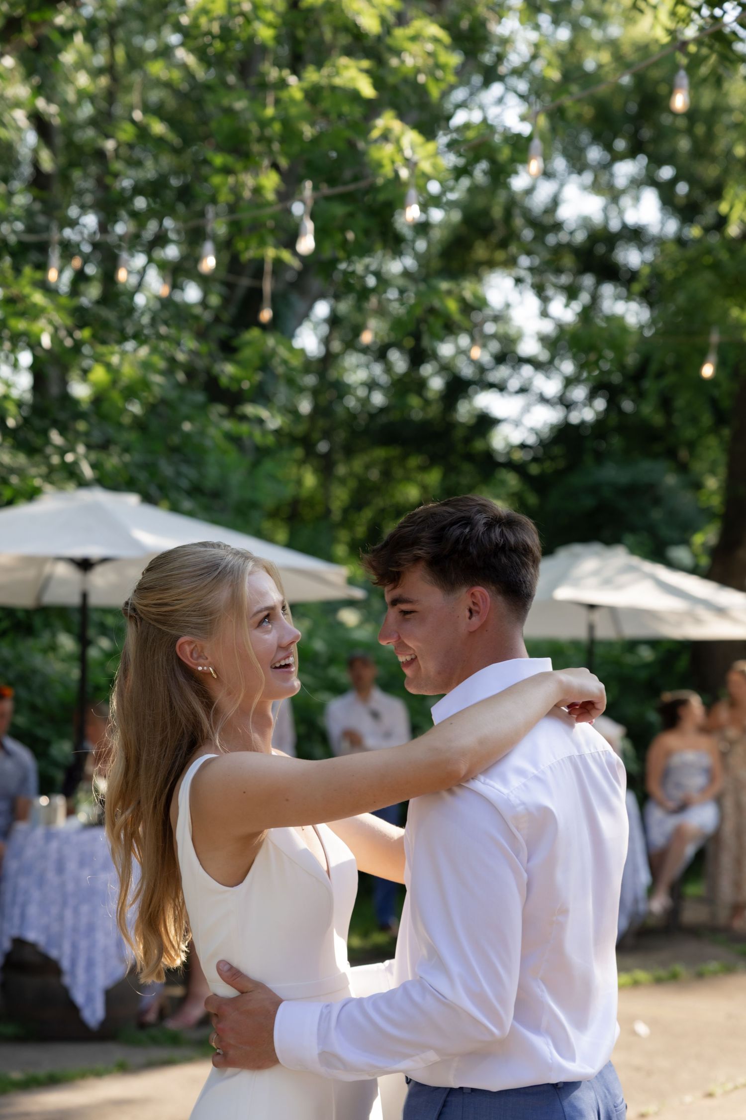 Newlyweds share their first dance at an outdoor garden reception under hanging lights and trees.