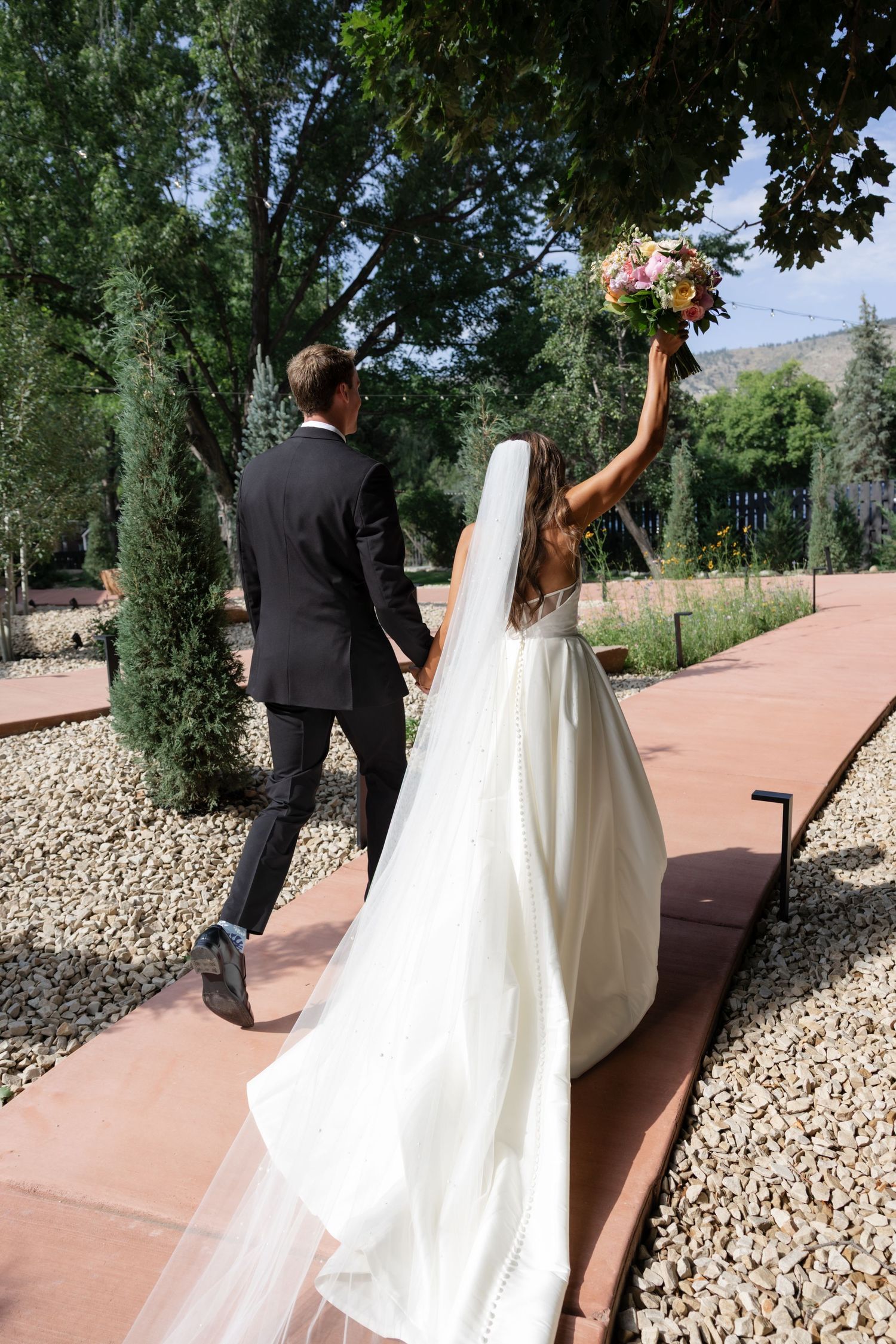 A bride in a white gown raises her bouquet while walking down a garden path with her partner.