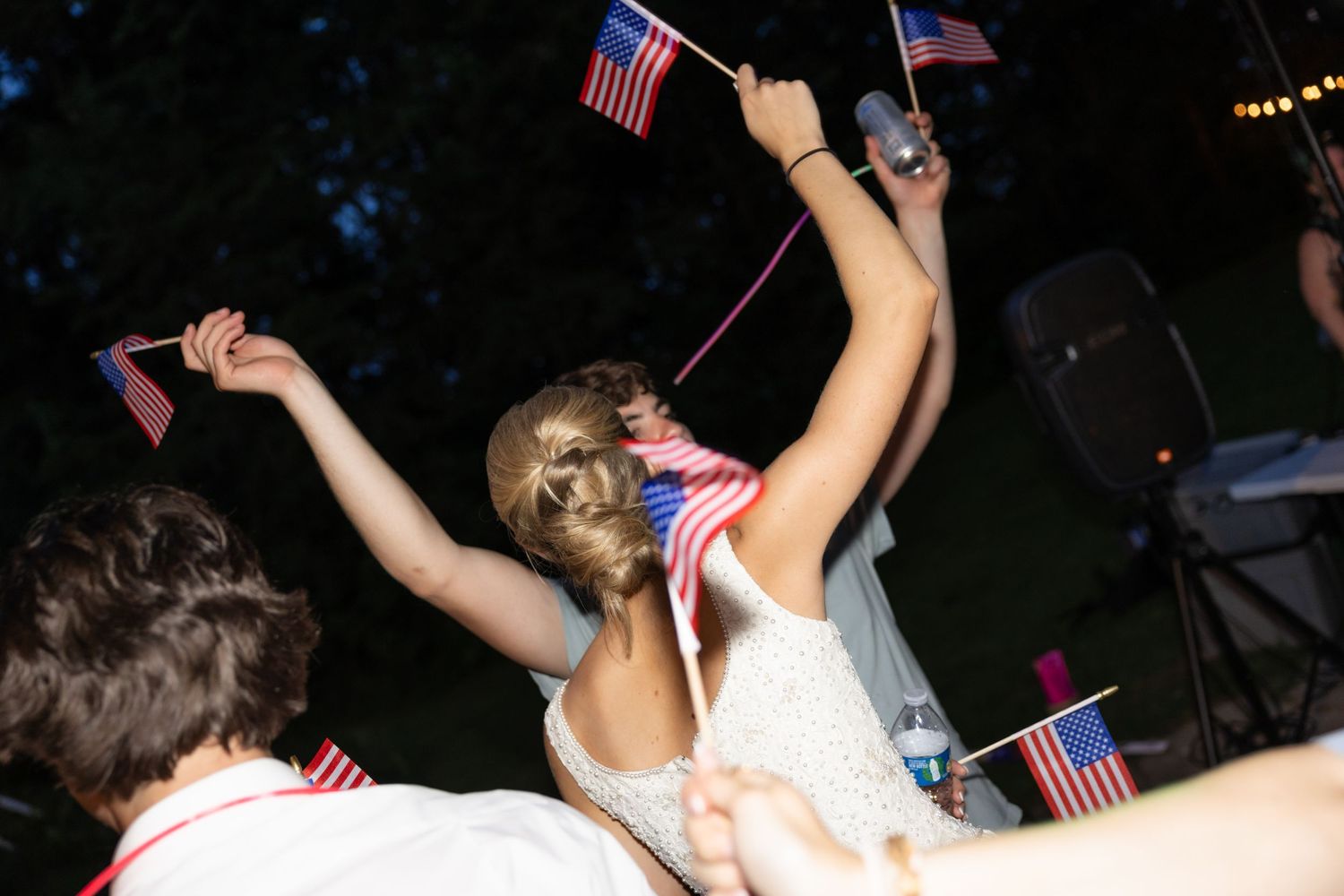 Crowd waving American flags at nighttime celebration with string lights overhead.