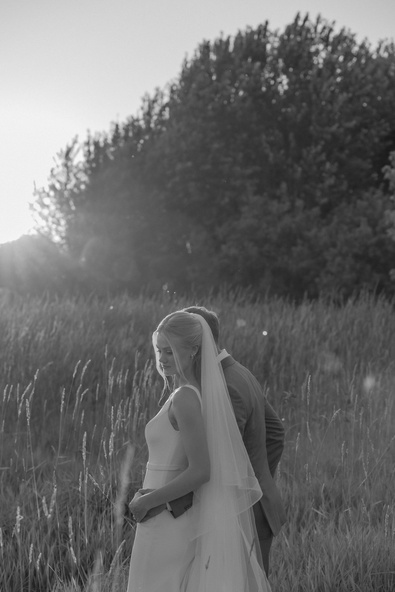 Black and white photo of newlyweds embracing in a field of tall grass at dusk.