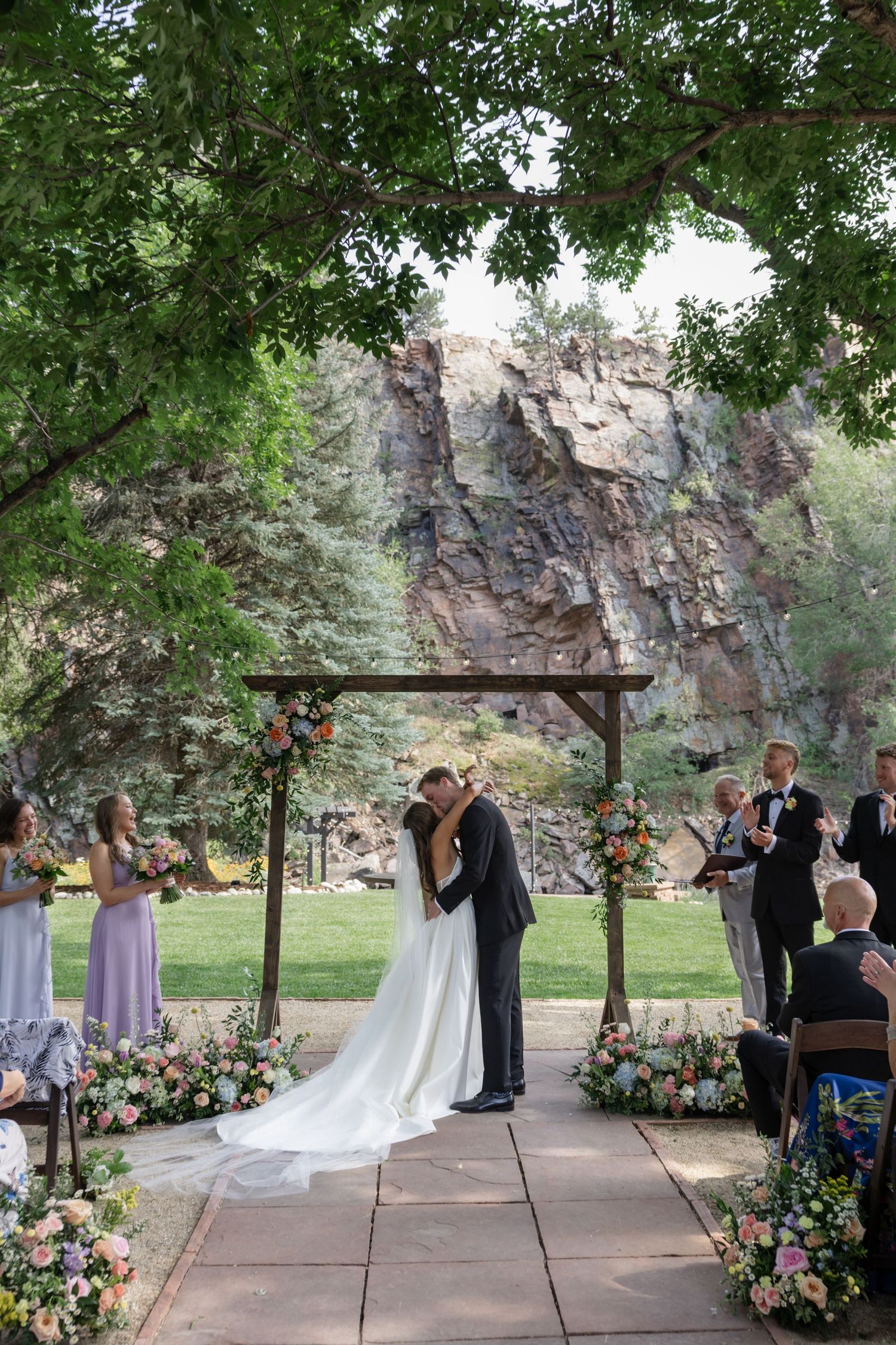A wedding ceremony takes place under a wooden arch with floral arrangements against a dramatic rocky cliff backdrop.