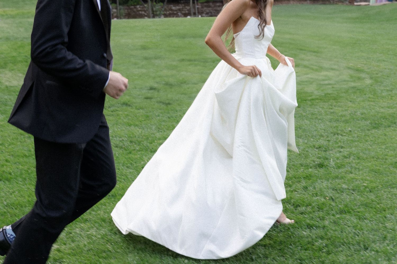 A couple in wedding attire dances playfully on a green lawn during their special day.