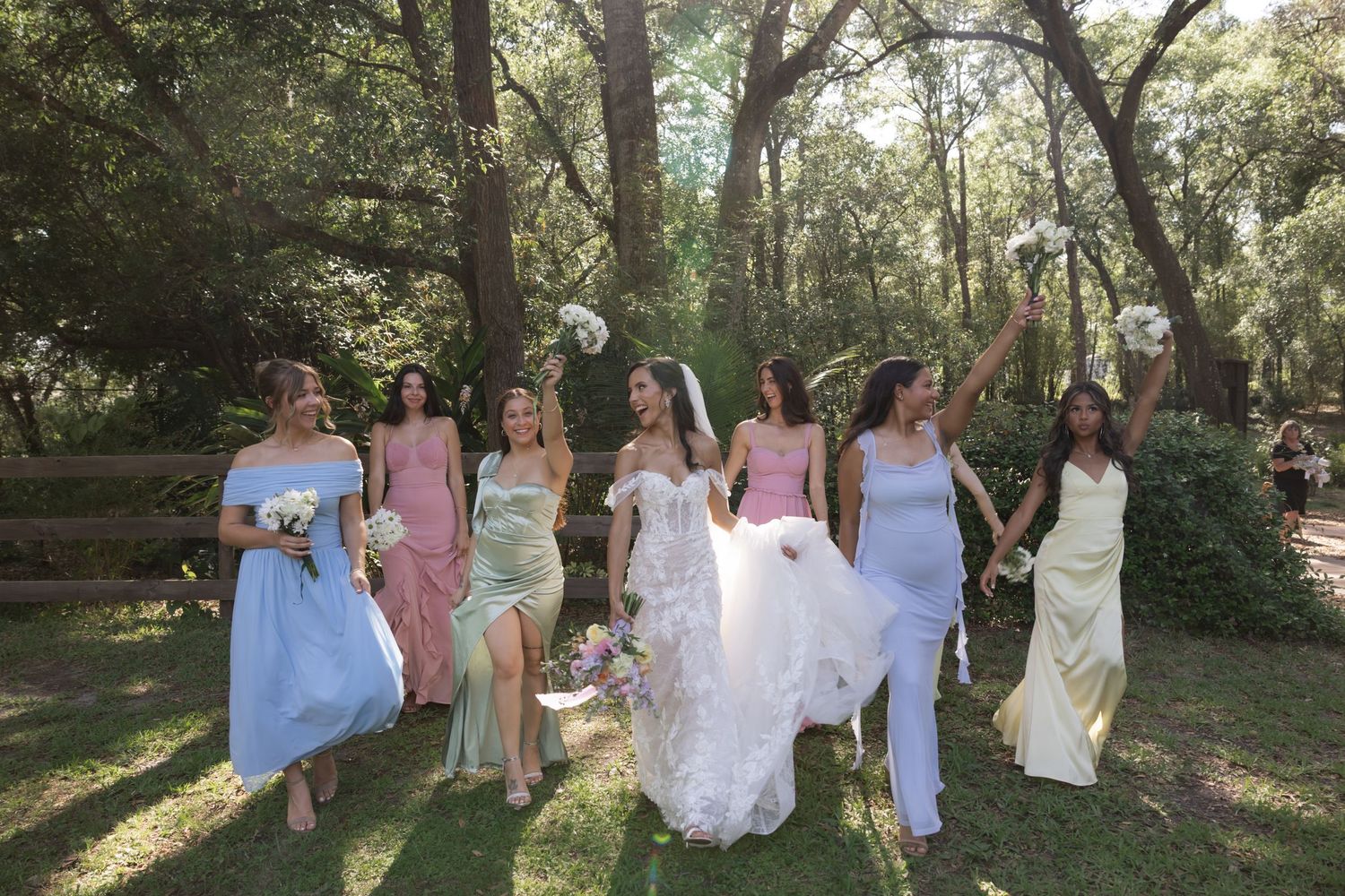Bridal party walking through a sunlit garden wearing pastel dresses and holding bouquets.