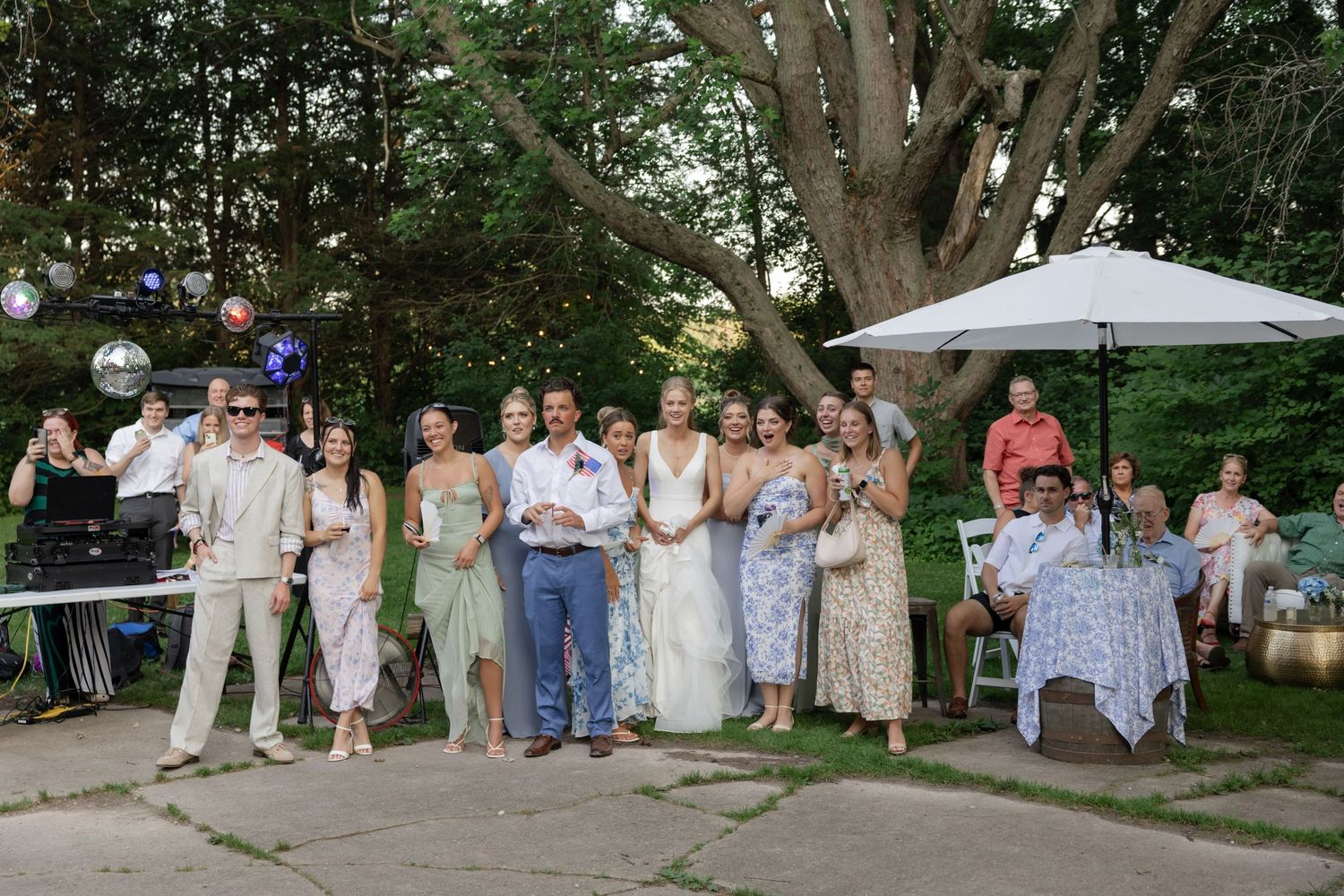 Wedding party and guests gathered outdoors for group photos under large trees.