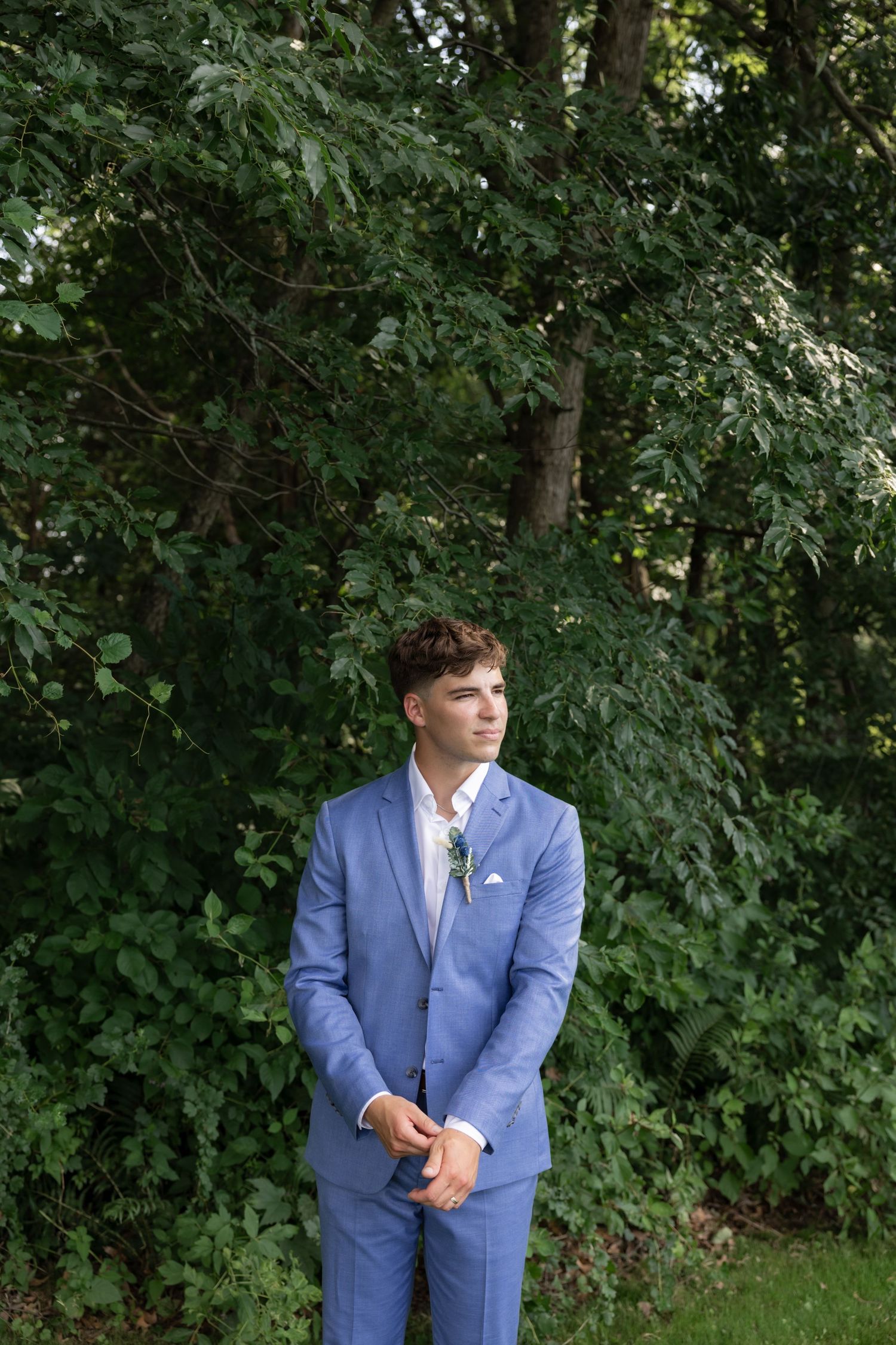 Wedding party member in a blue suit stands in front of lush green trees.