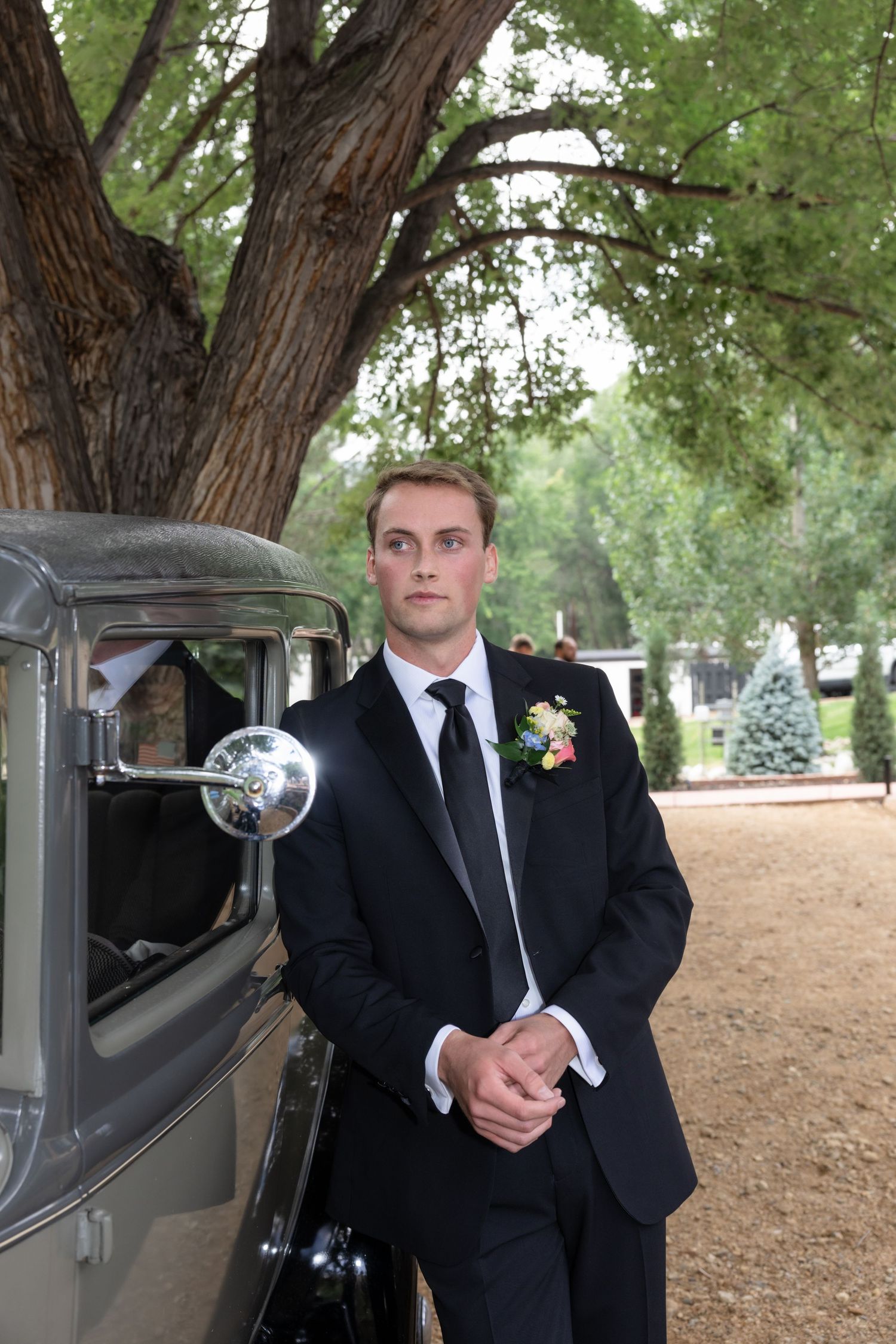 Groom poses with classic car outdoors wearing black tuxedo.