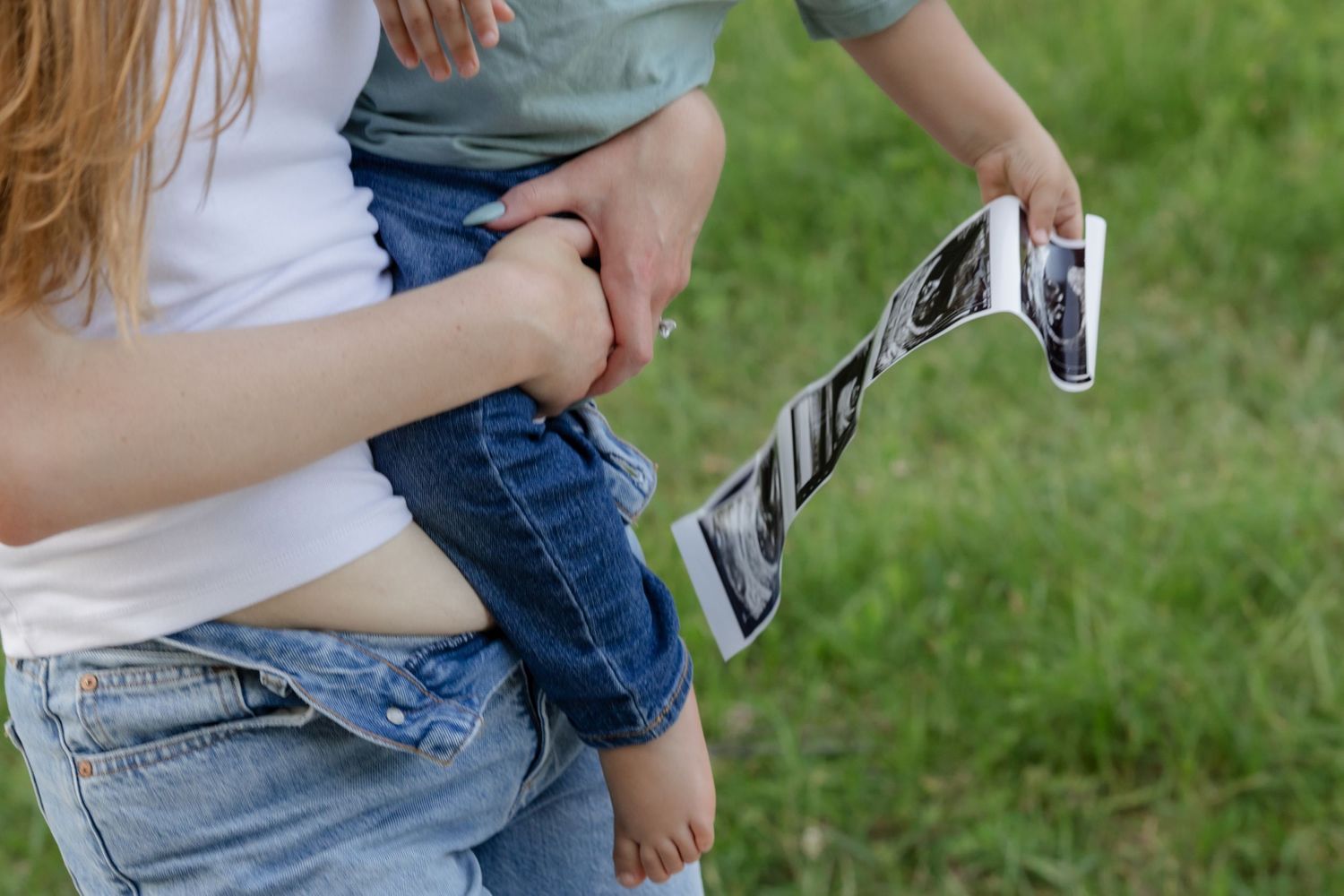 Individual wearing denim and holding ultrasound photos outdoors with a green lawn background.