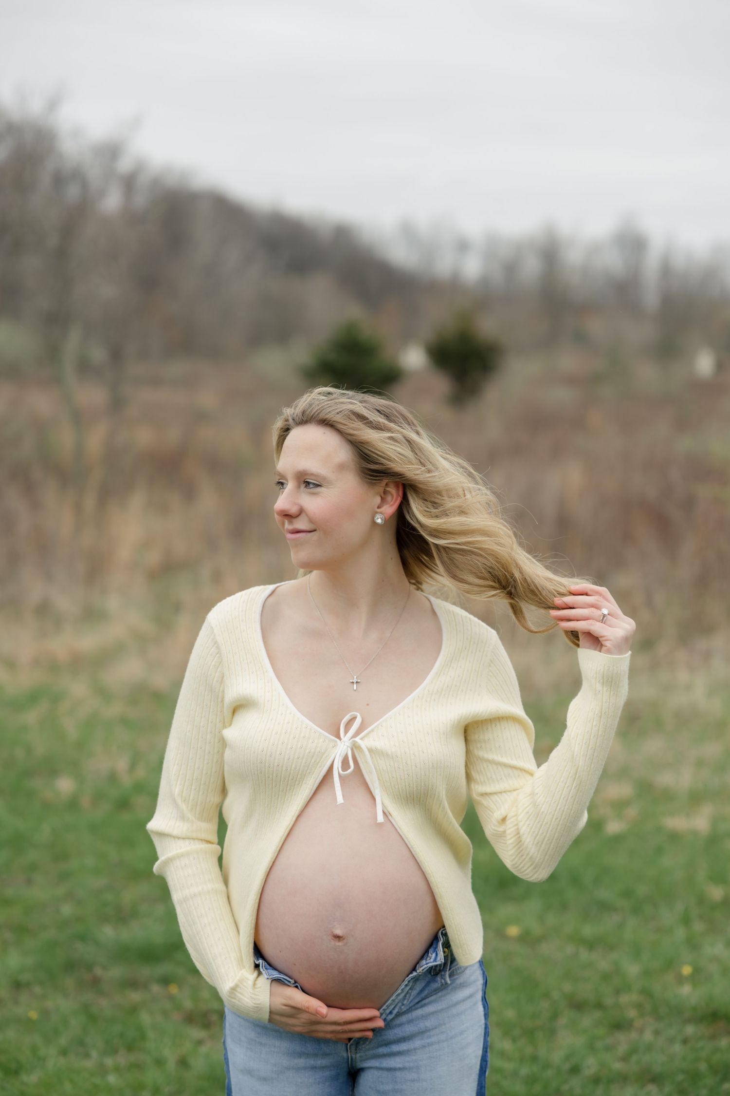 A pregnant woman in a pale yellow cardigan posing joyfully in a grassy meadow.