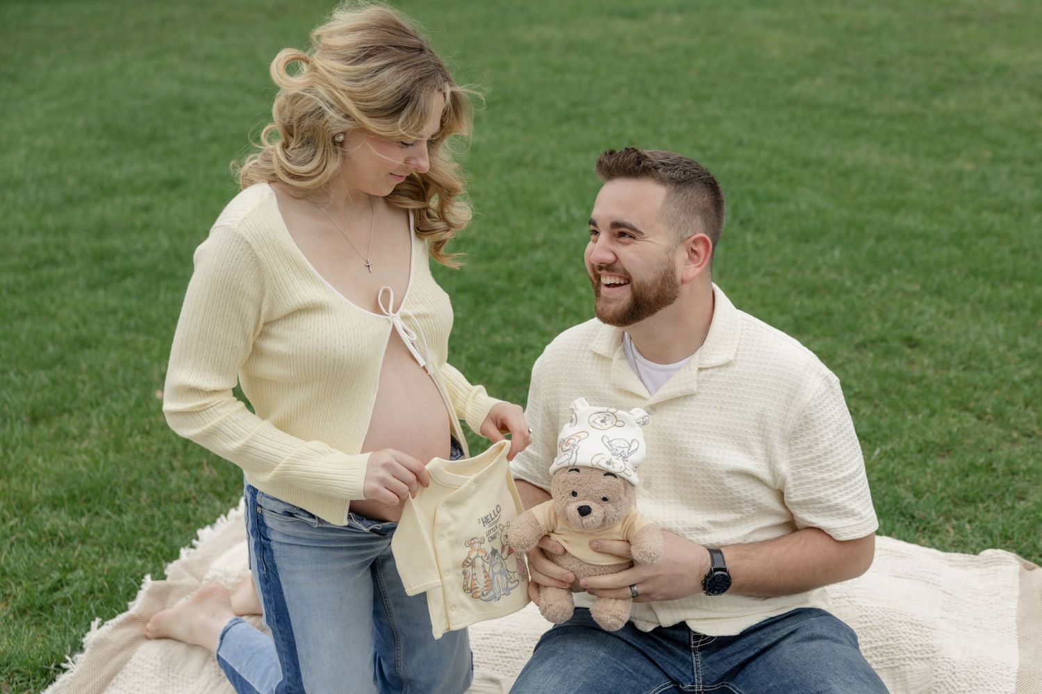 Expectant couple sitting on blanket outdoors holding teddy bear during maternity photoshoot.