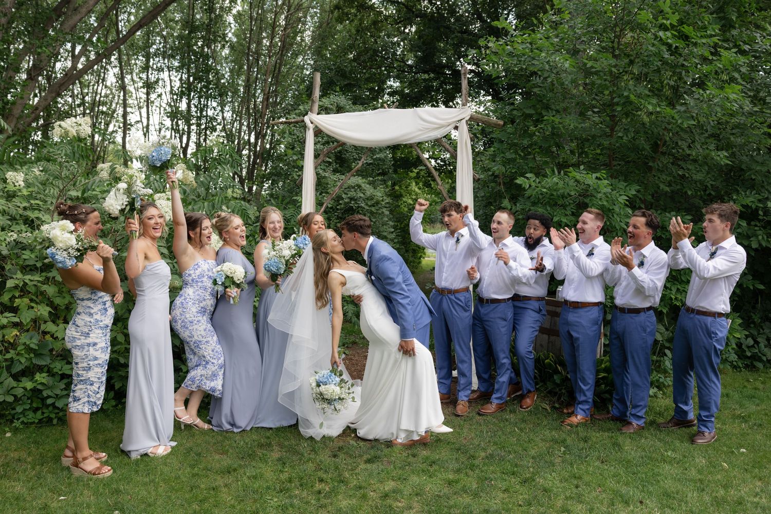 Large wedding party in blue and gray attire posing together outdoors against green foliage.