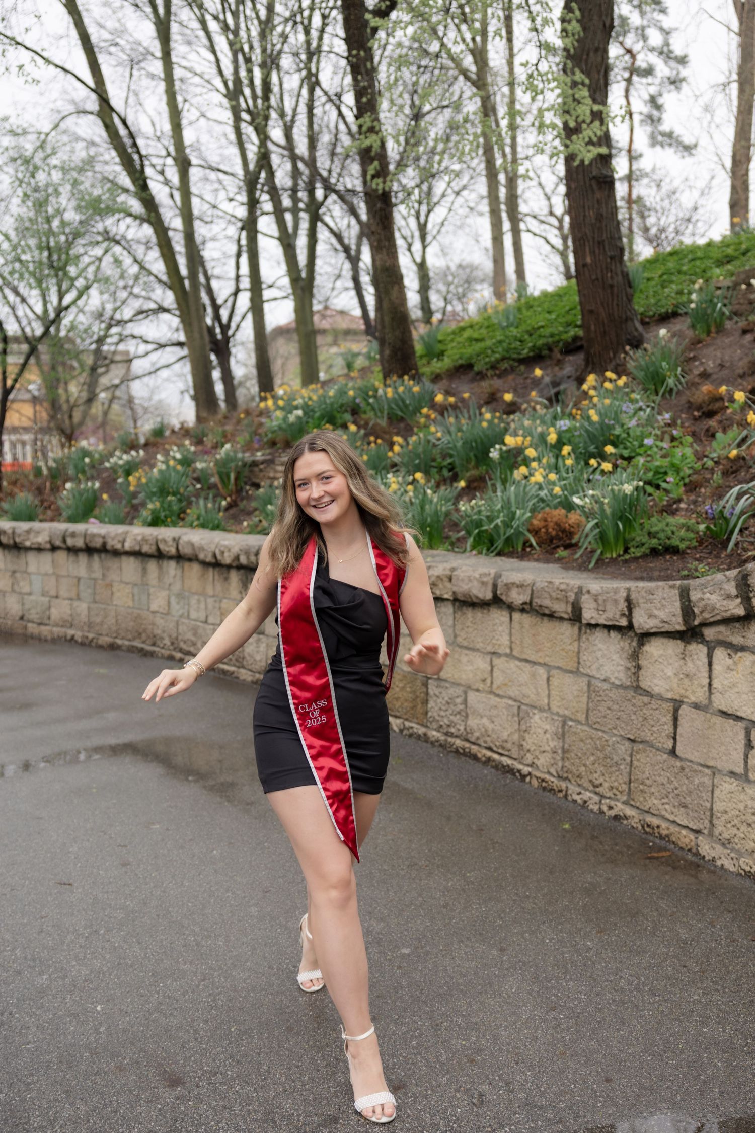 A person in graduation attire poses playfully along a stone retaining wall lined with spring flowers.