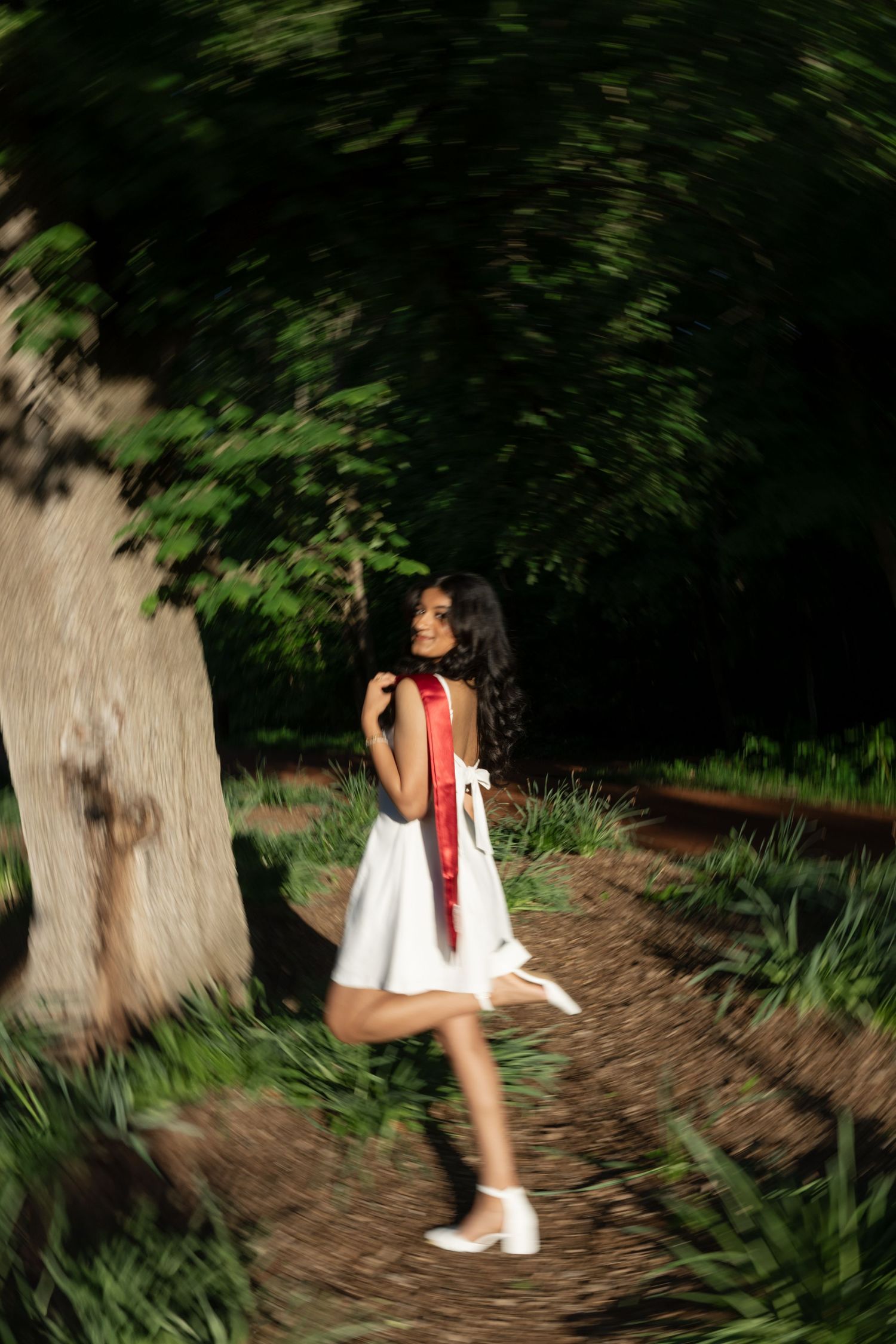 A person in white dress and red sash walks along a wooded path in dappled sunlight.