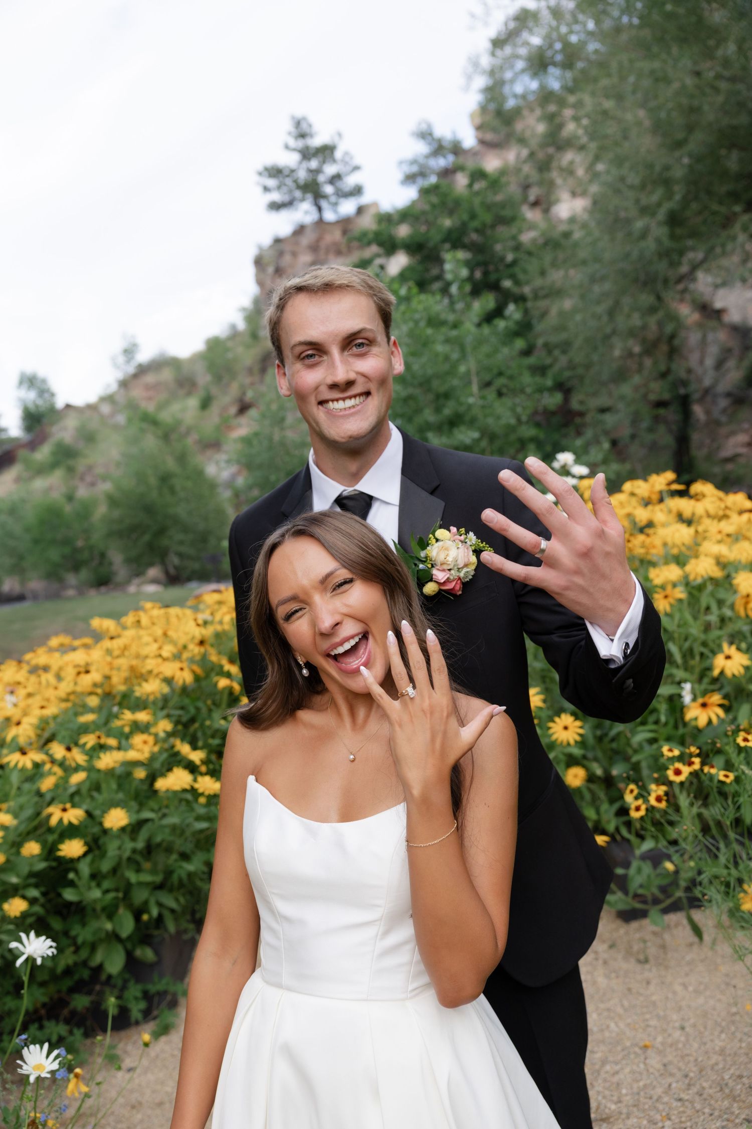 Playful wedding couple poses joyfully among yellow flowers in an outdoor garden setting.