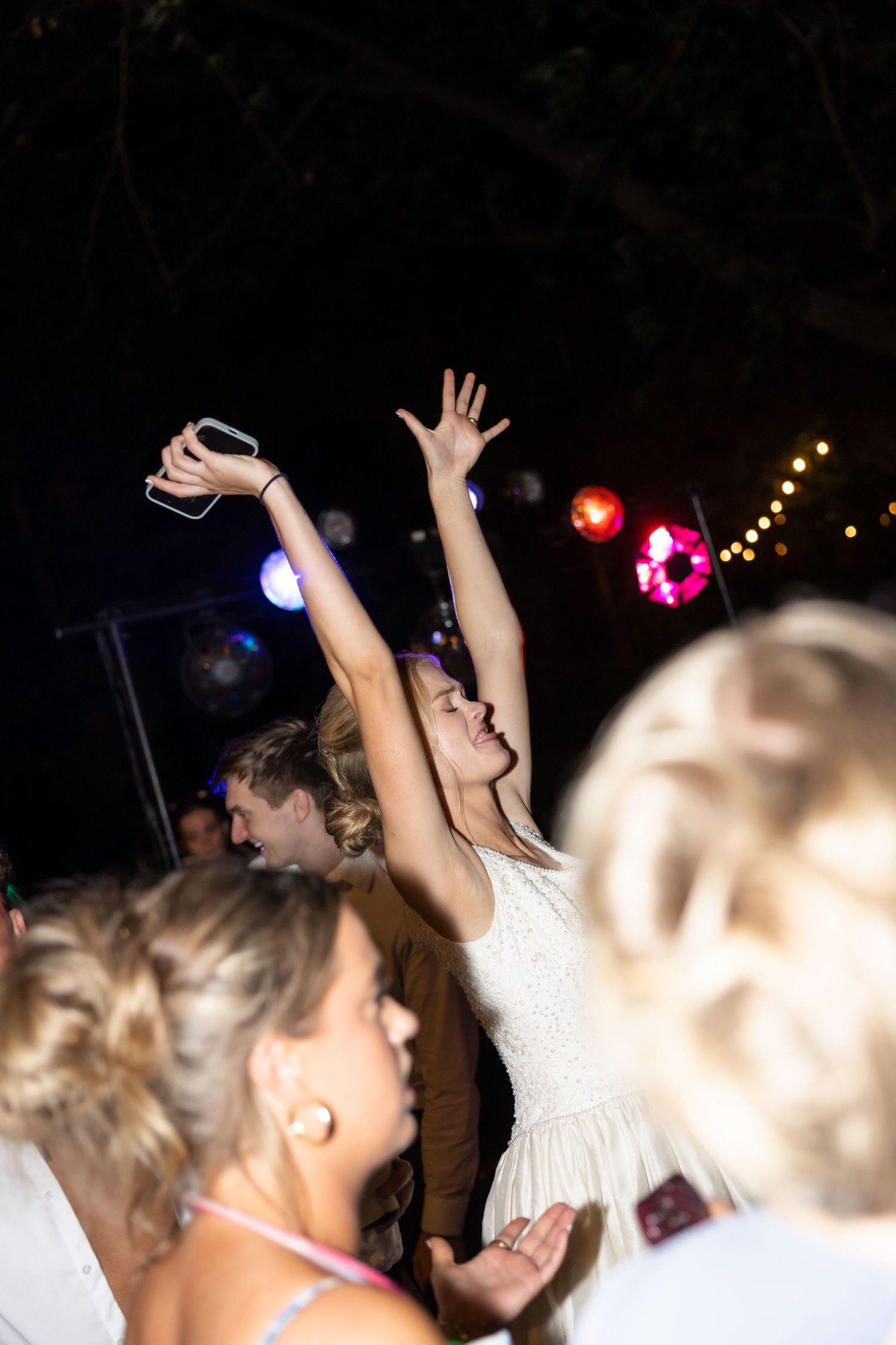 Crowd with raised hands dancing under string lights at an outdoor party.