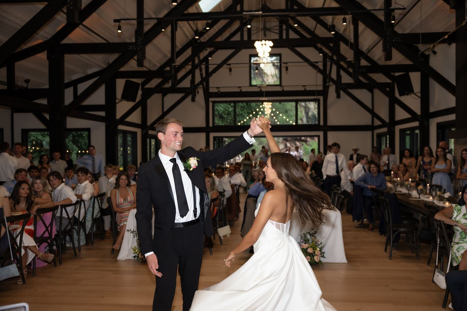 Wedding couple spins during romantic dance in rustic reception hall with guests watching.
