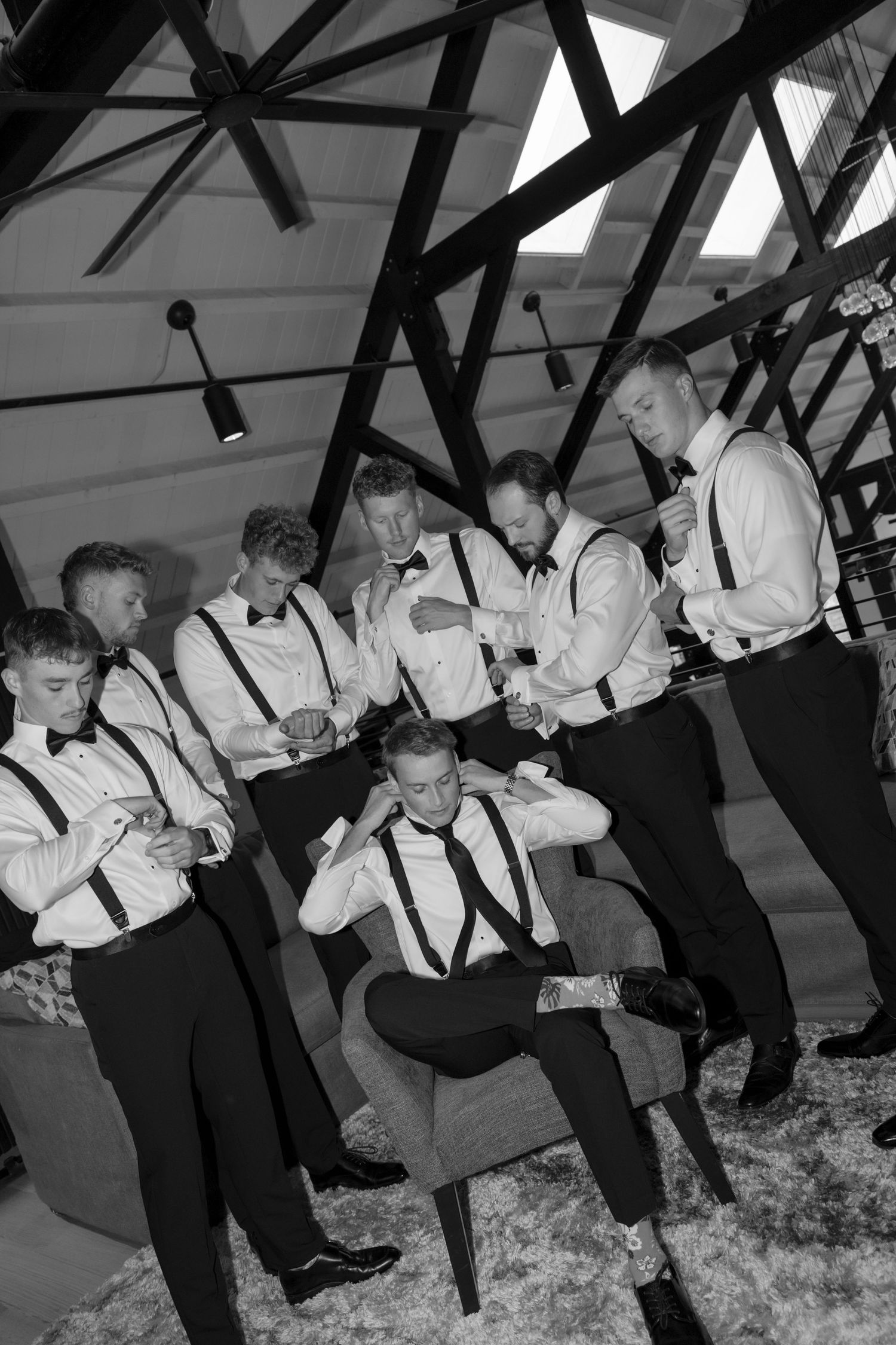 Groomsmen in black and white attire gather in a rustic venue with exposed wooden beams overhead.