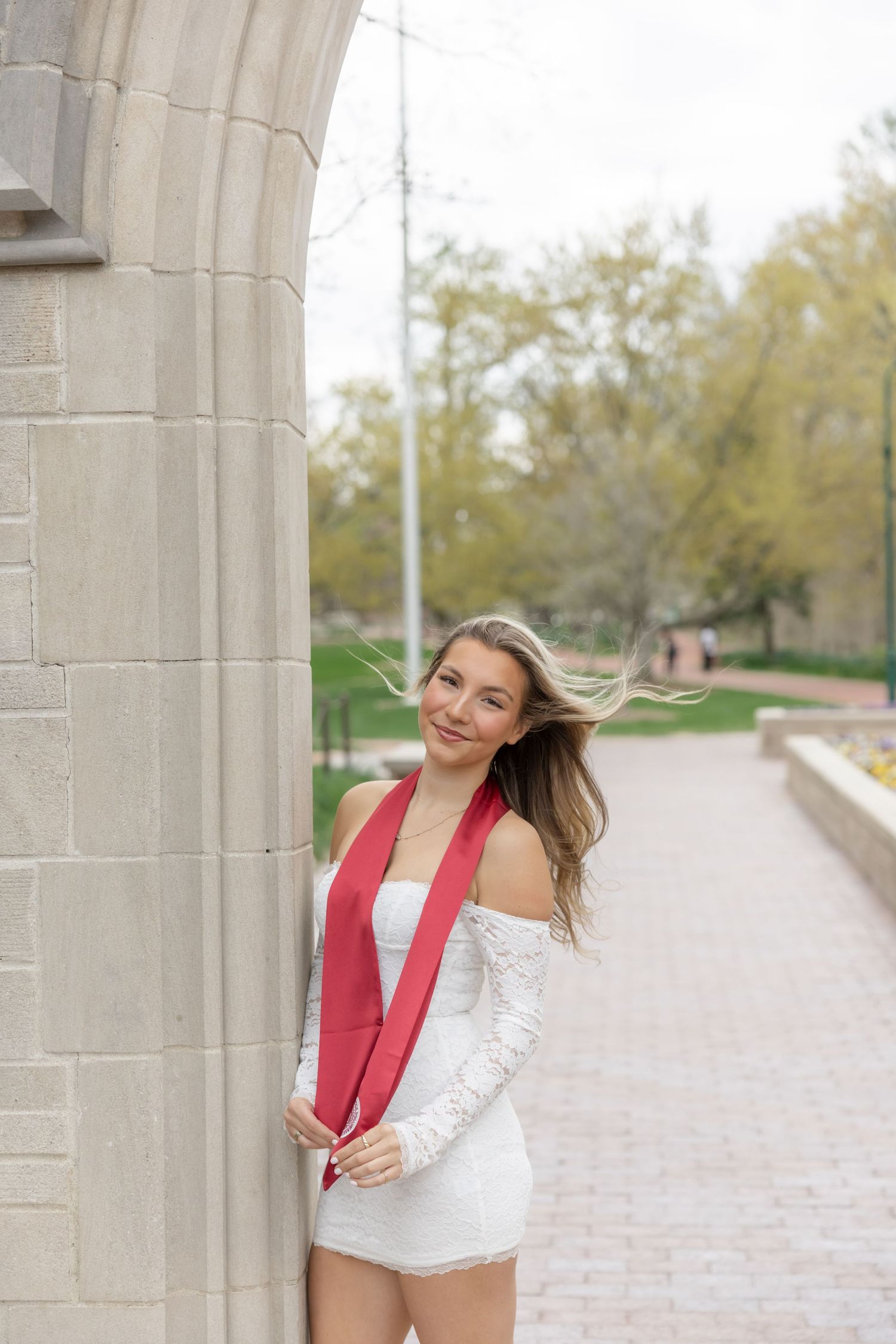 Someone wearing a white top and red stole poses playfully between stone columns on a brick pathway.