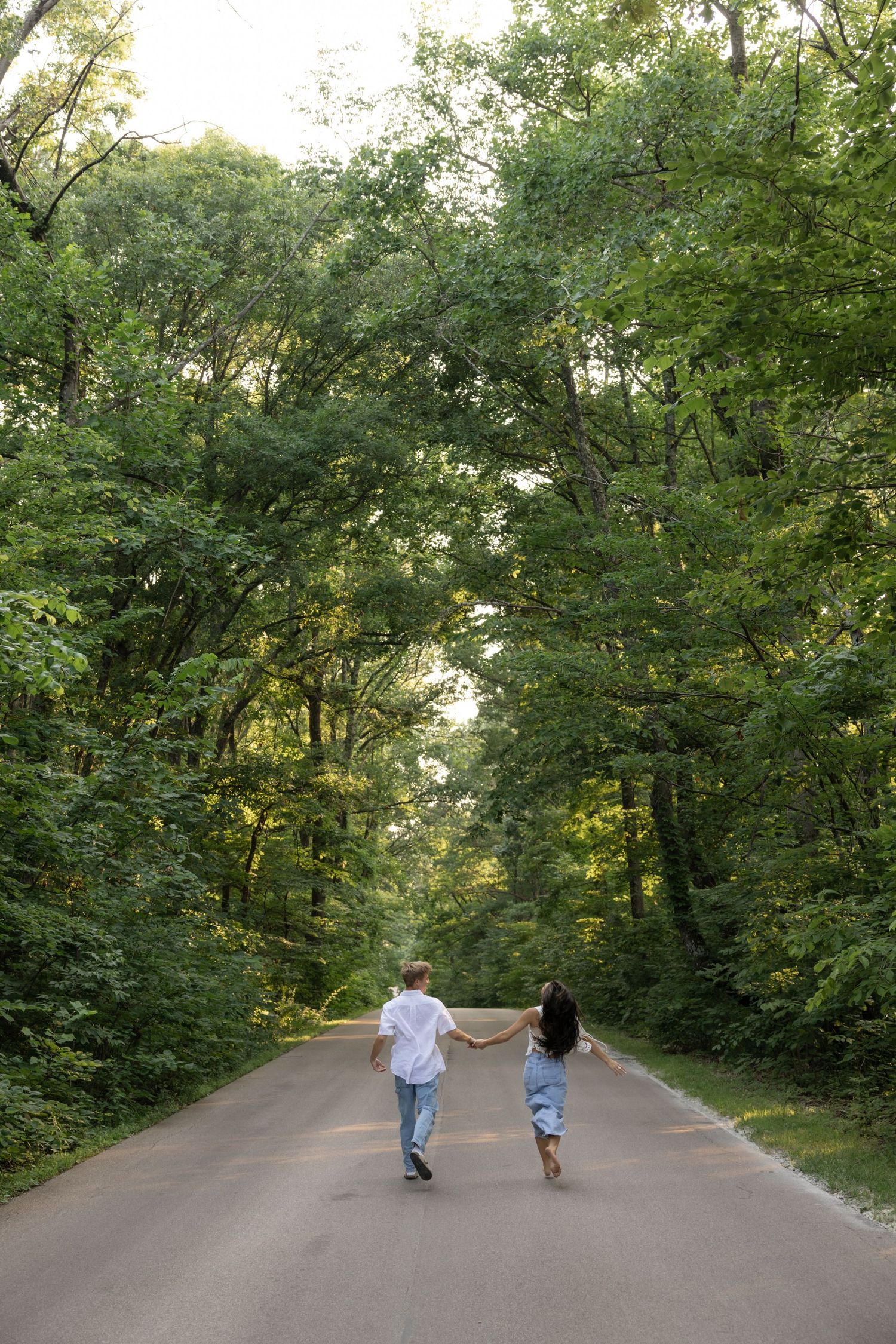Two people holding hands run down a tree-lined road together on a summer day.