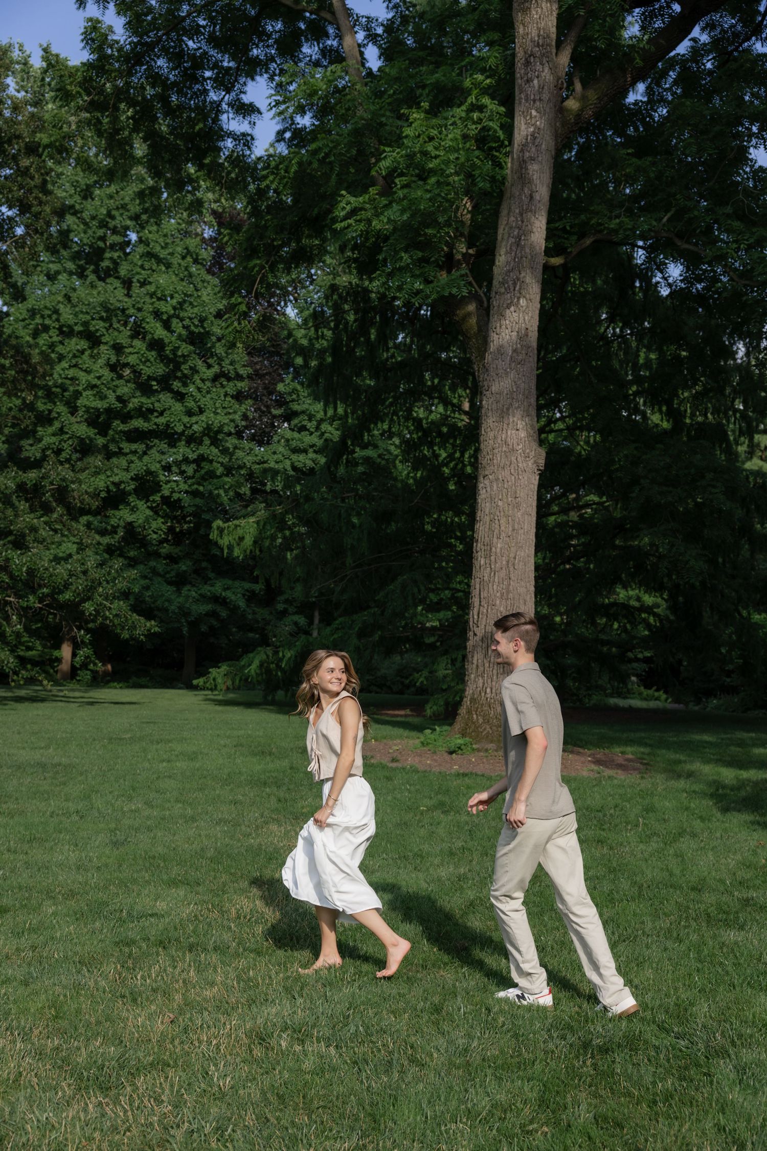 A couple playfully runs through a lush green park with tall trees in the background.