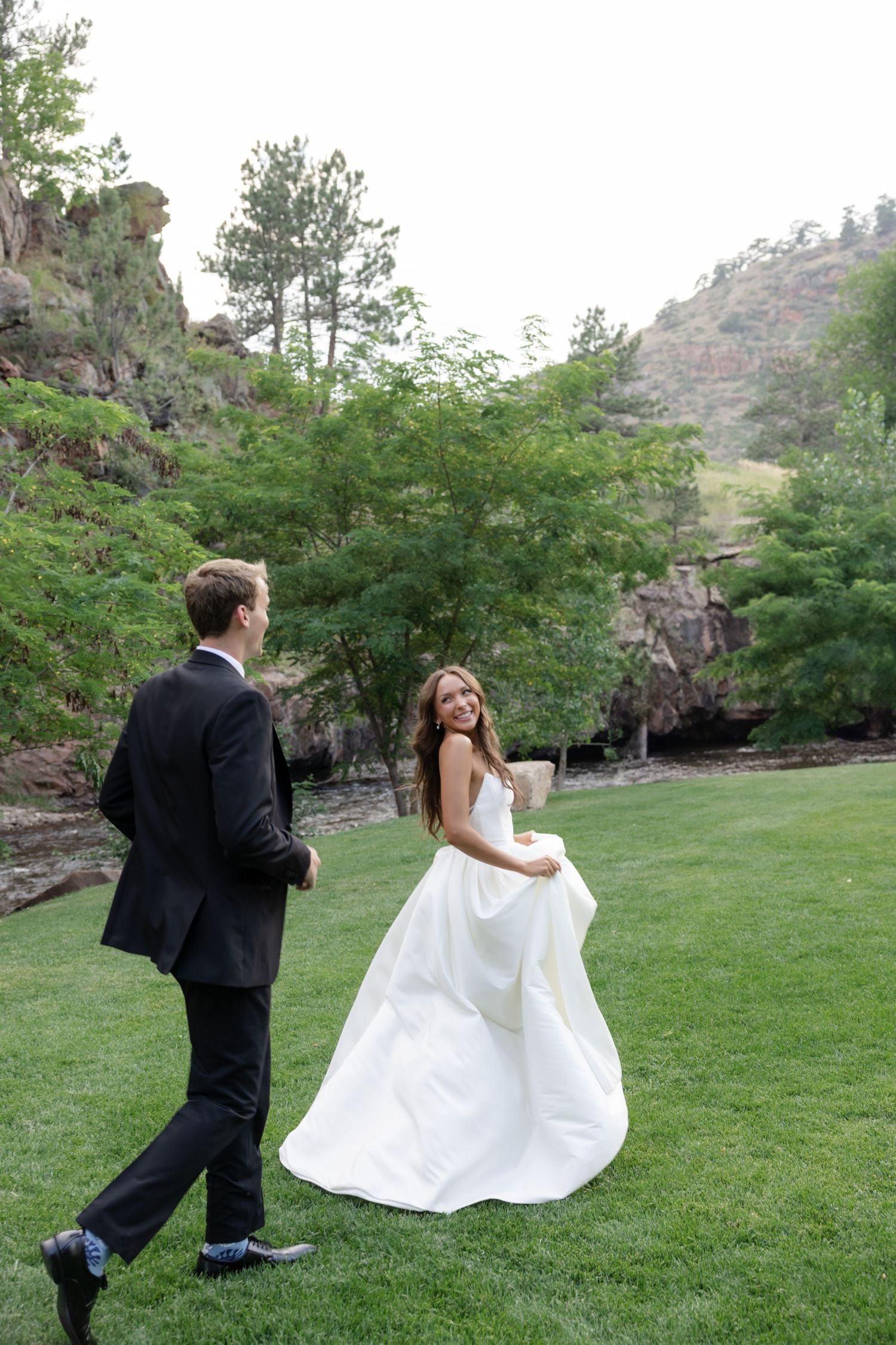 A wedding couple plays and dances together on a lush lawn with rocky cliffs in the background.