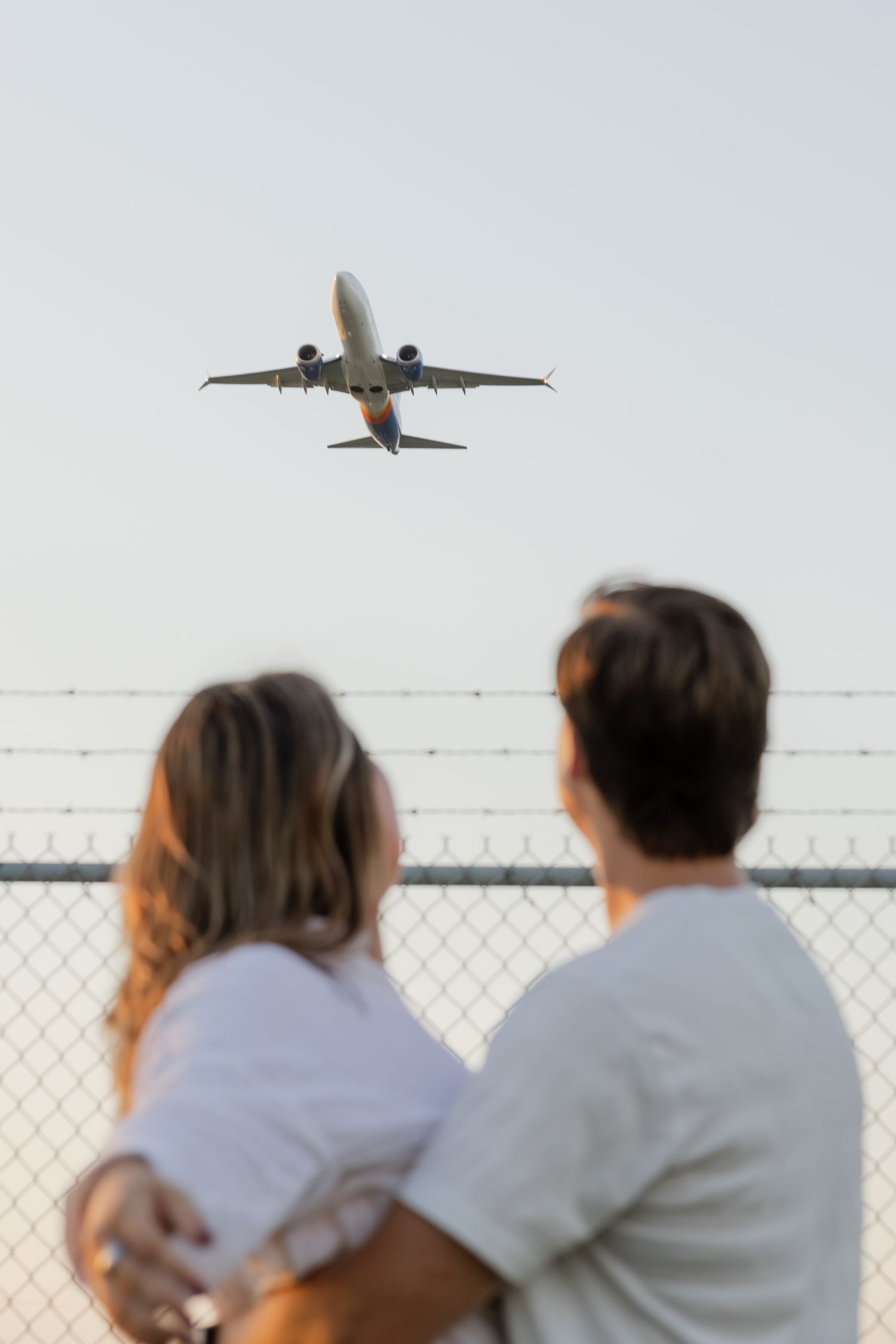 A couple watches commercial airplanes flying overhead near an airport fence.