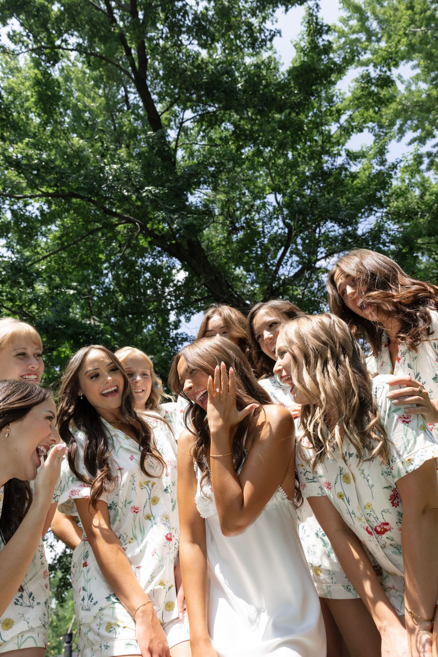 Group wearing floral and white outfits posing together outdoors under green trees.