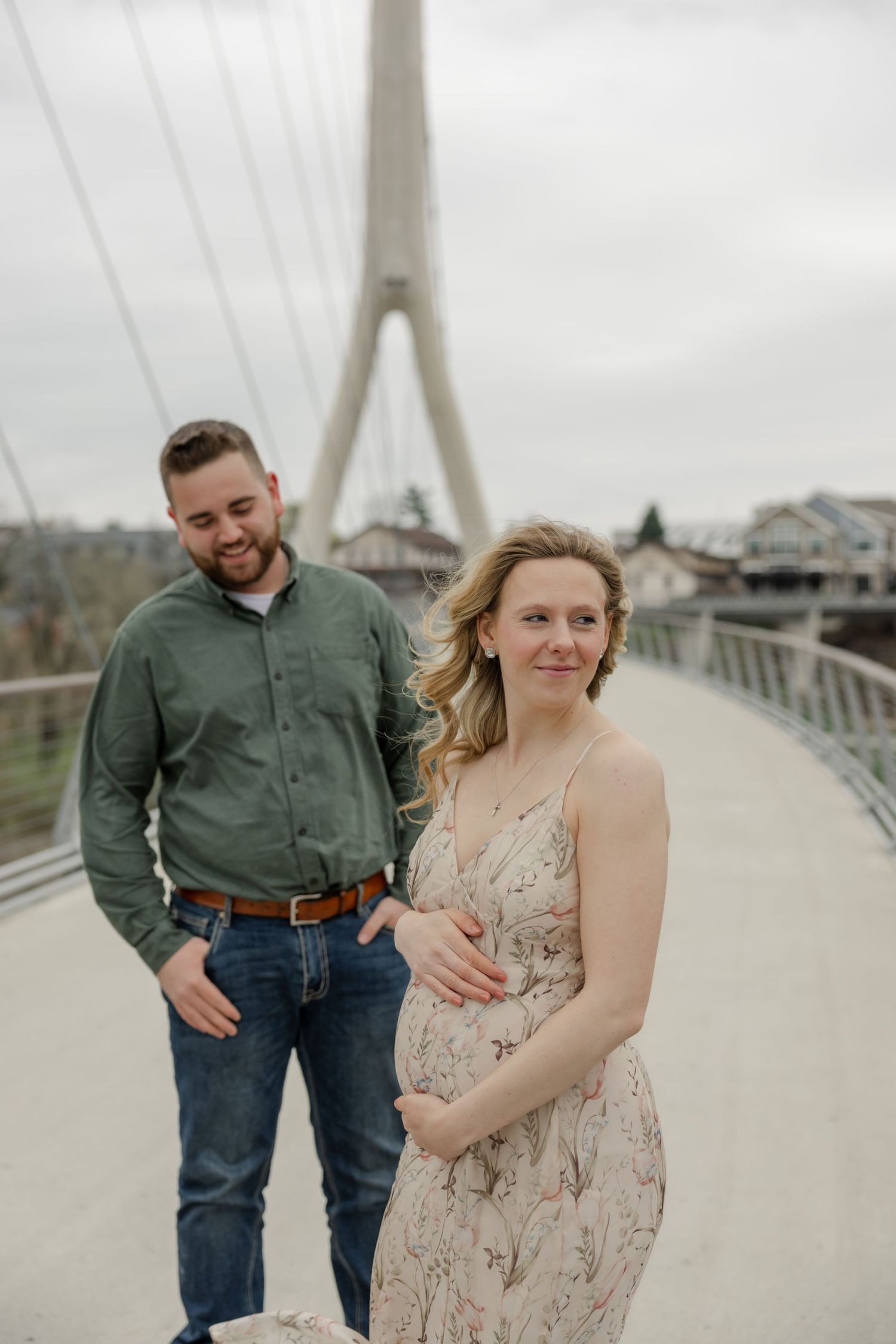 A couple poses for maternity photos on a bridge with cloudy skies in the background.