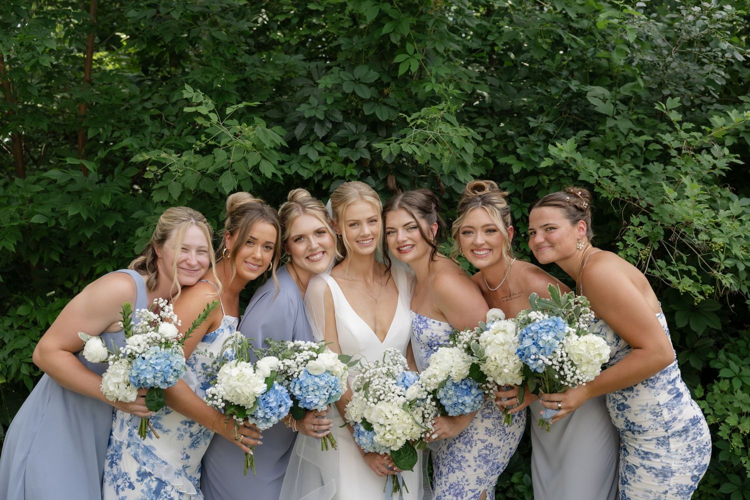 A group in elegant dresses holds blue and white floral bouquets at an outdoor wedding celebration.