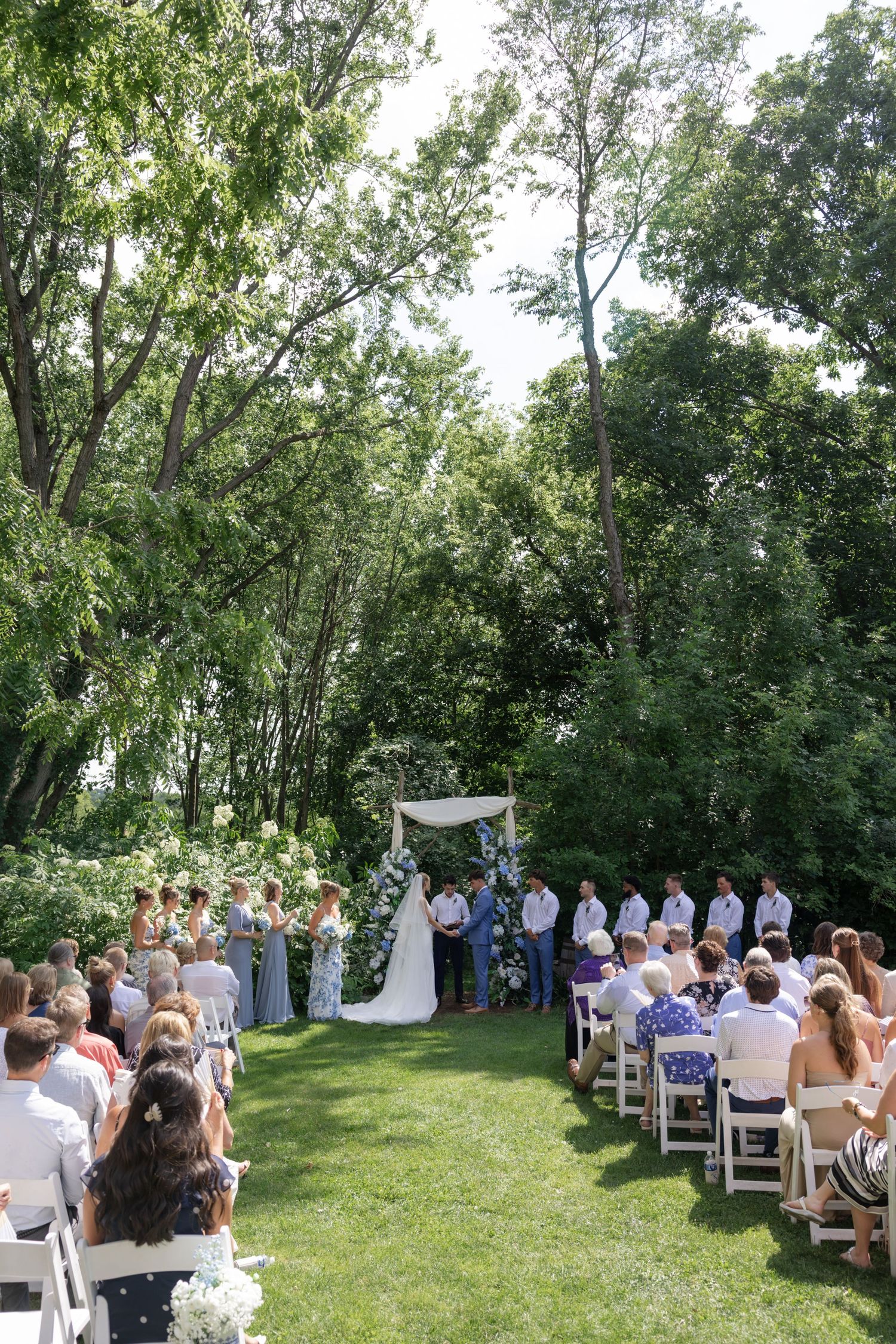 Wide view of an outdoor wedding ceremony with guests seated in white chairs under a canopy of trees.