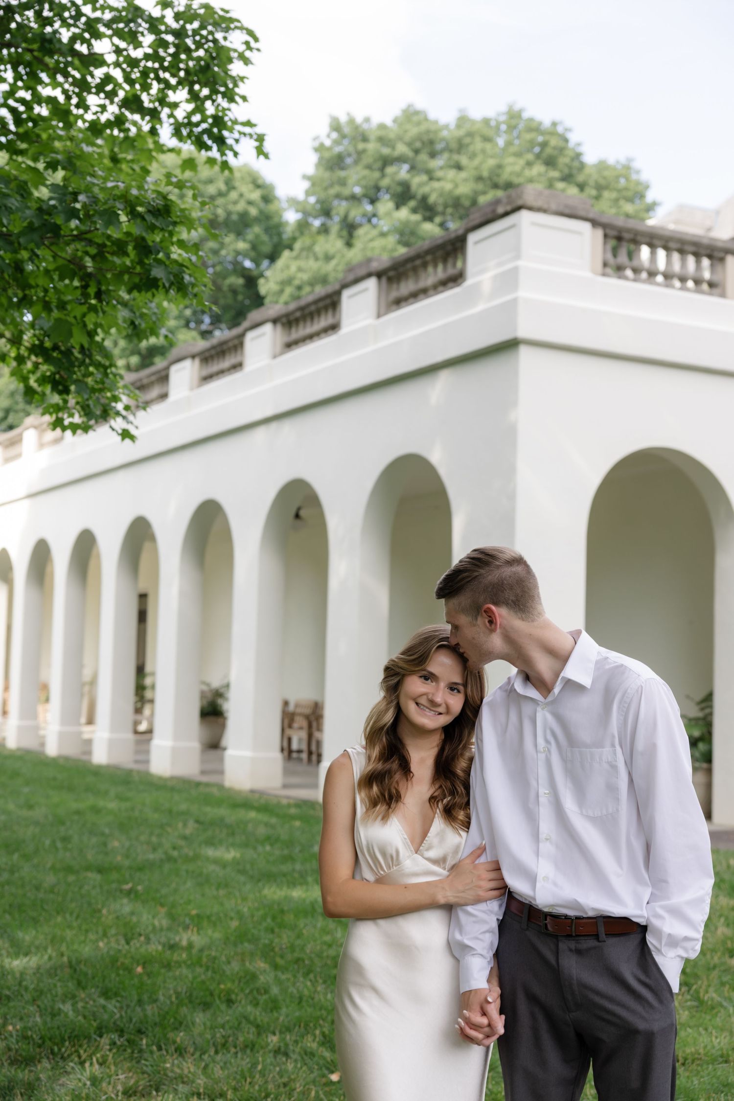 A couple shares an intimate moment in front of a white columned building with arched architecture.