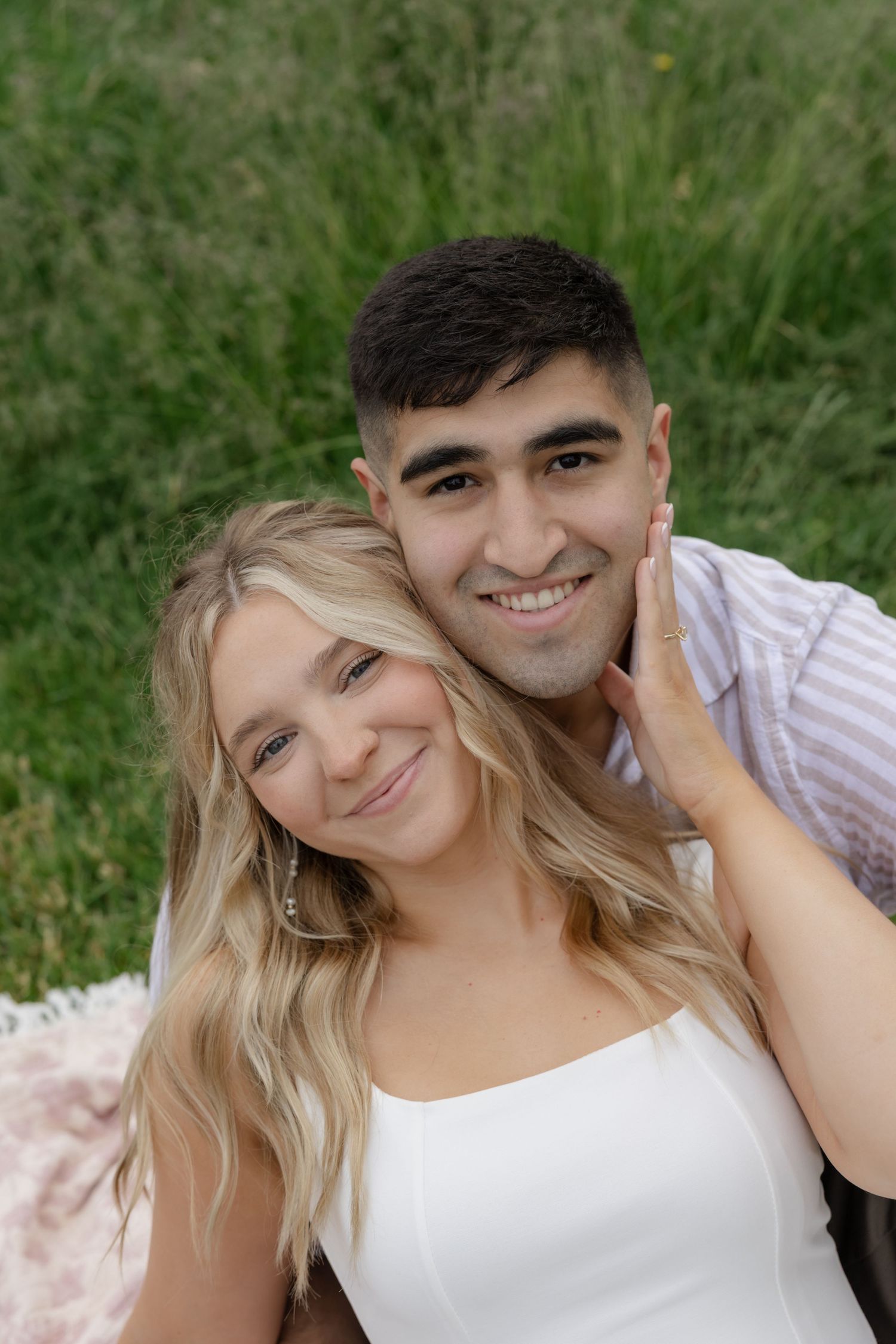 A couple smiles and embraces while sitting together in a sunlit grassy field.
