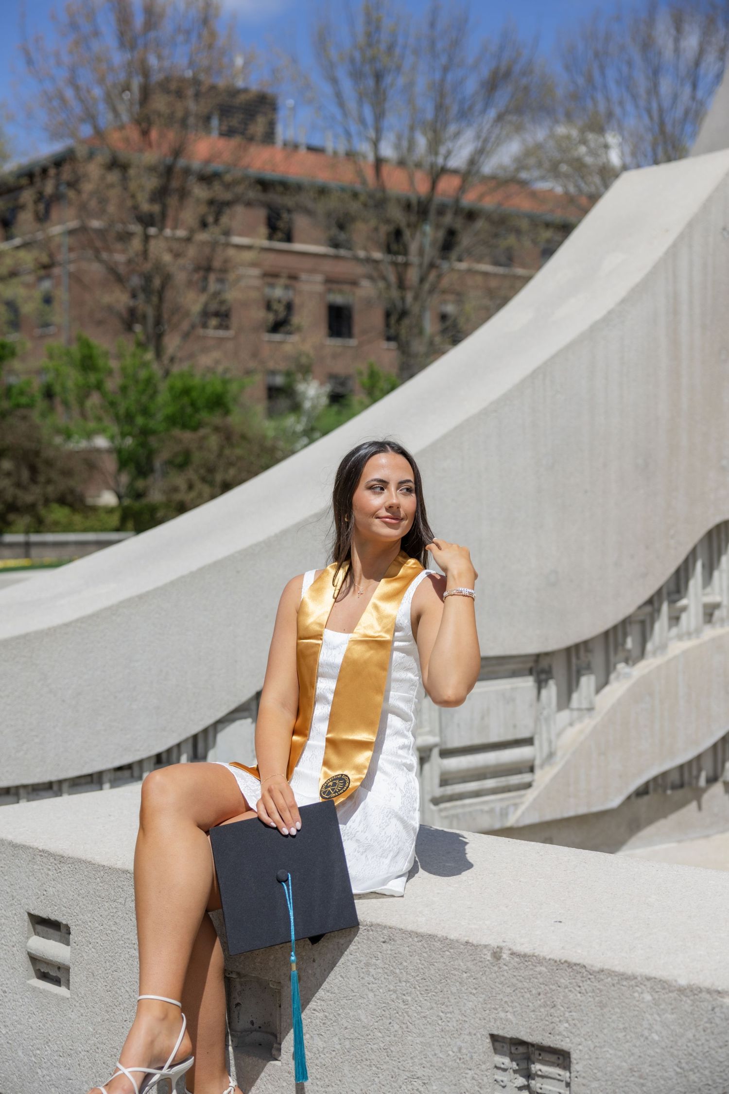 Graduate in white dress and gold sash poses on modern concrete steps with graduation cap.