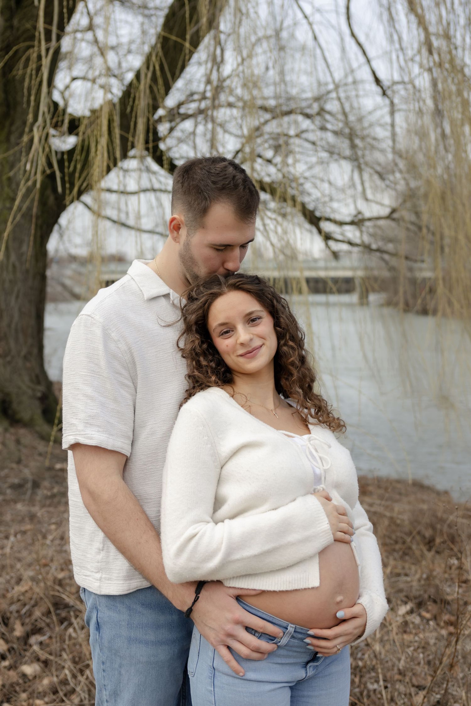 Couple embracing outdoors near willow trees in casual attire during maternity photoshoot.