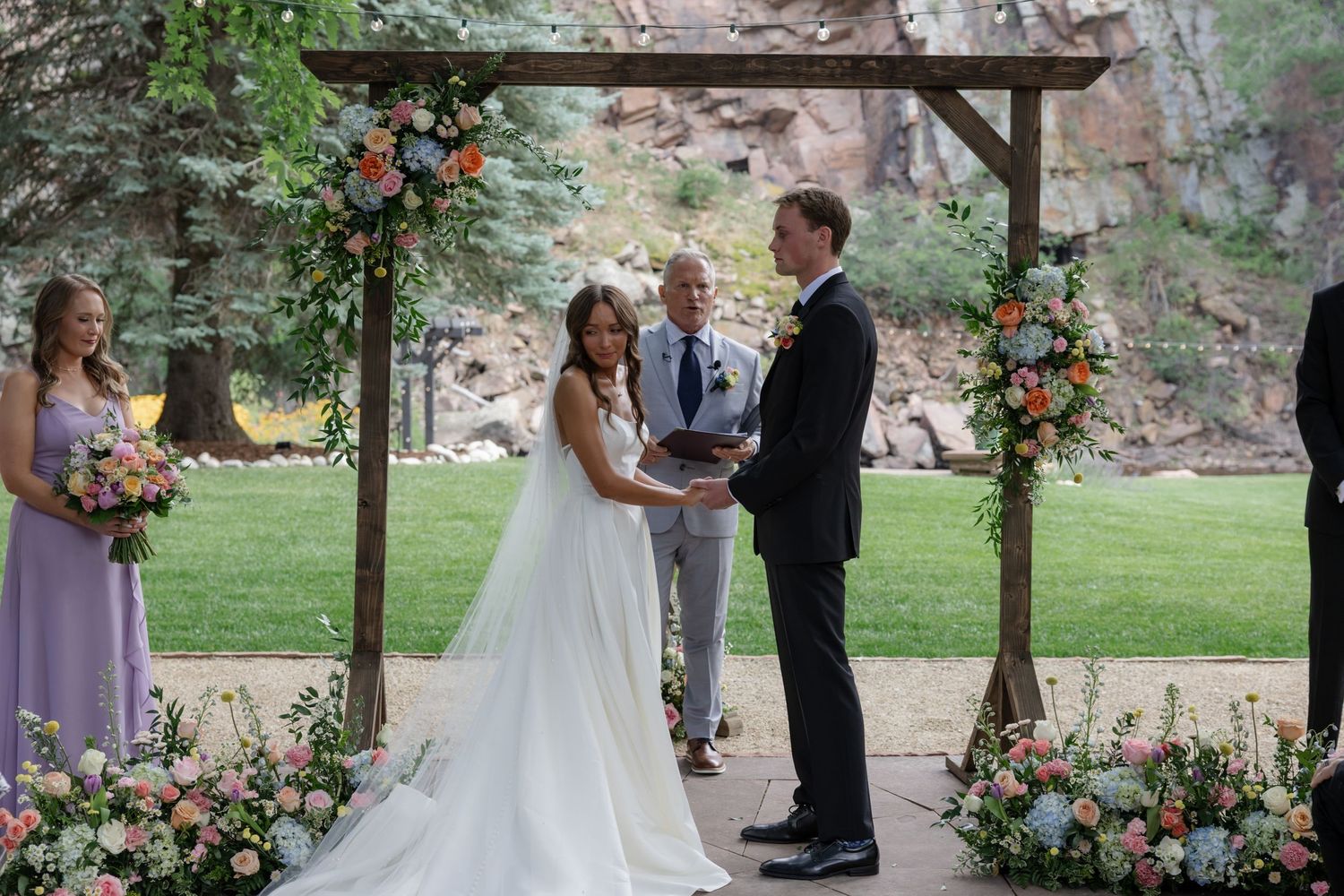 Wedding ceremony site with wooden pergola decorated with colorful flowers against natural rock formations.