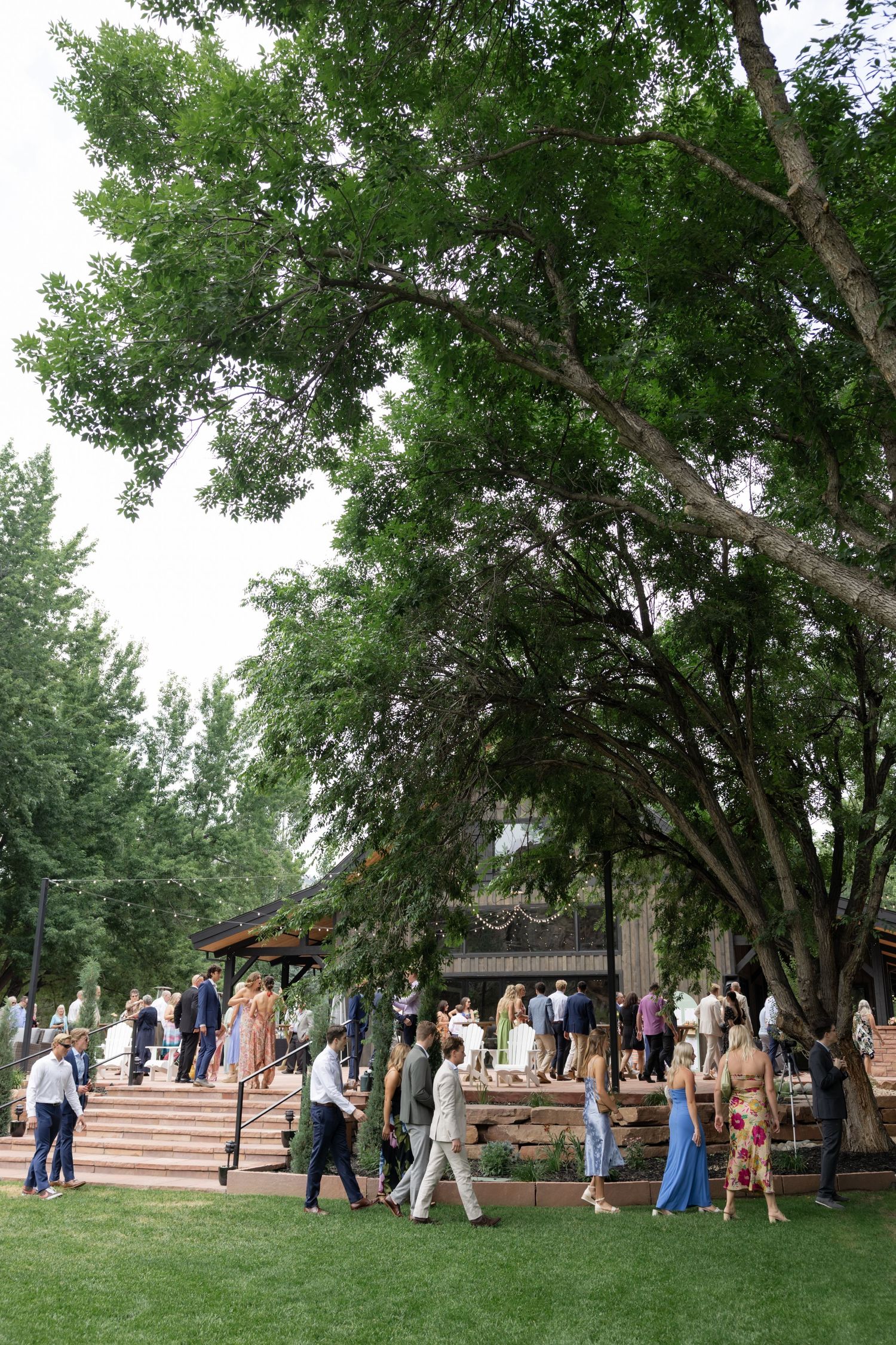 Wedding guests gather outside under large shade trees at an outdoor venue.