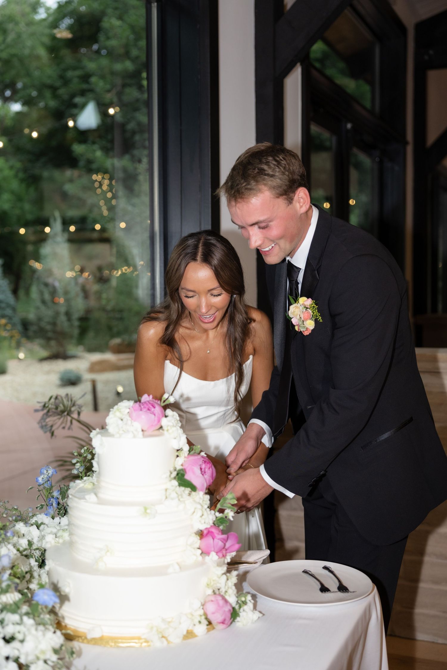 Traditional cake cutting moment at wedding reception with white tiered cake and pink flowers.