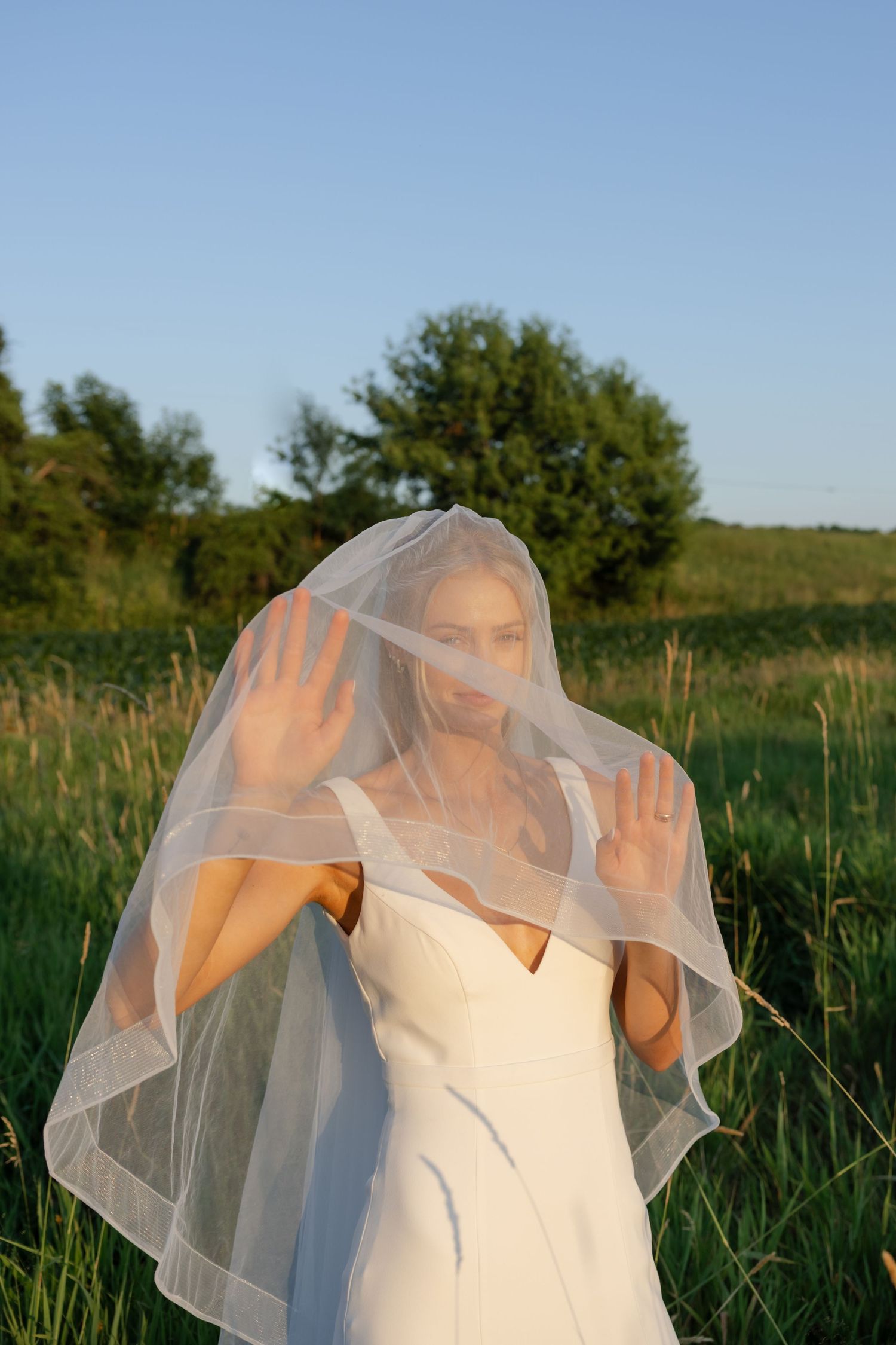 Bride adjusts her flowing veil in golden sunlight while standing in a green field.