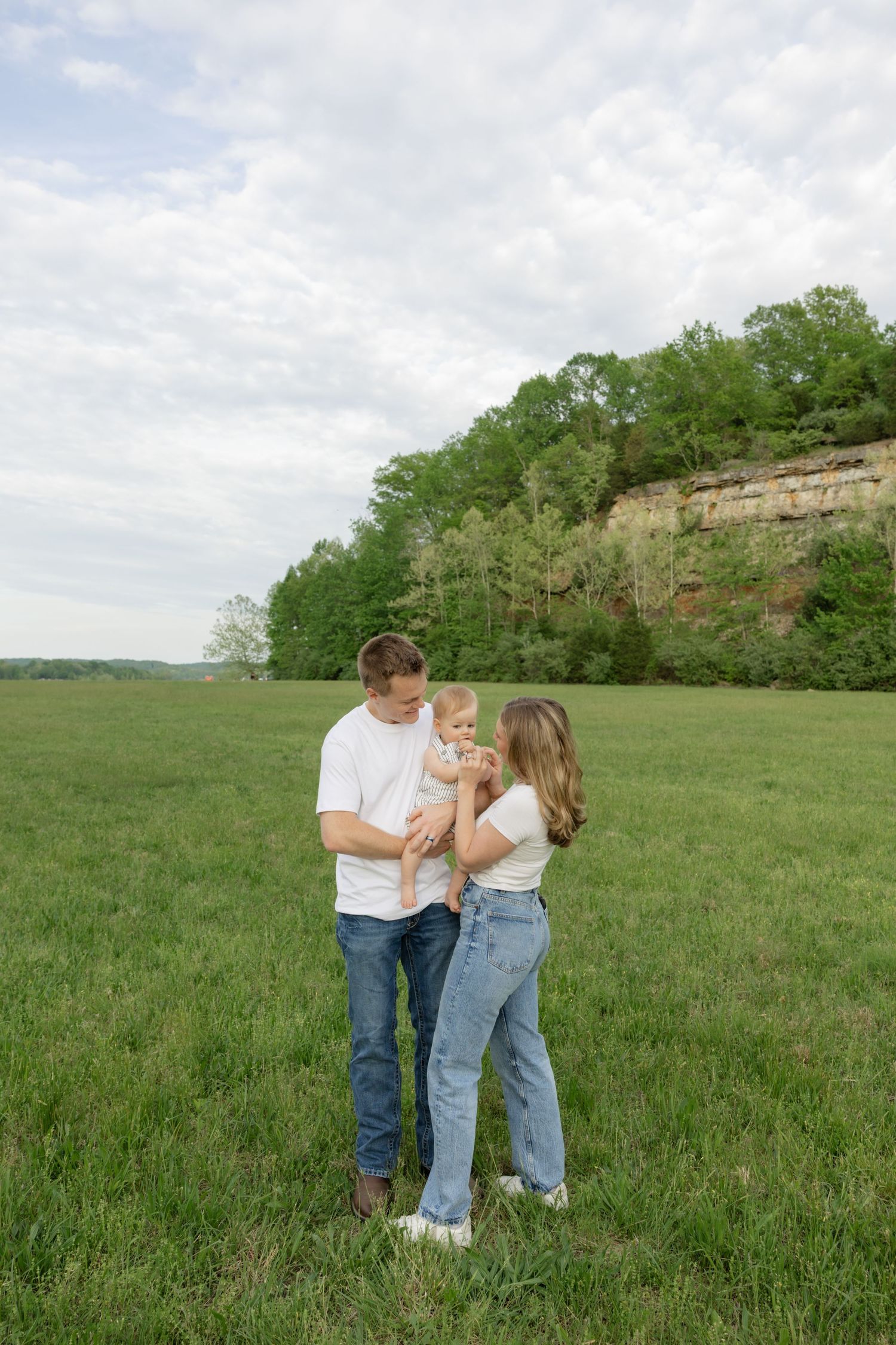 Family shares tender moments together in a sunlit grassy field near rocky cliffs.