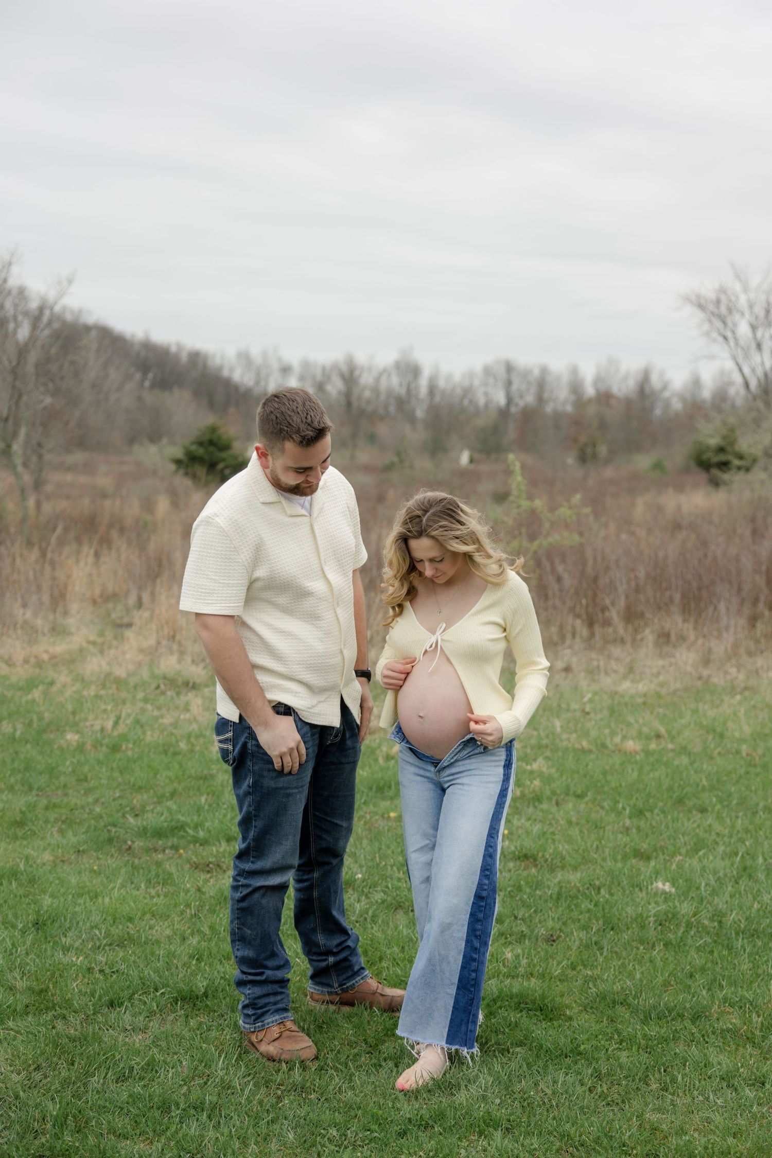 Couple in cream and denim outfits having candid moment during outdoor maternity shoot.