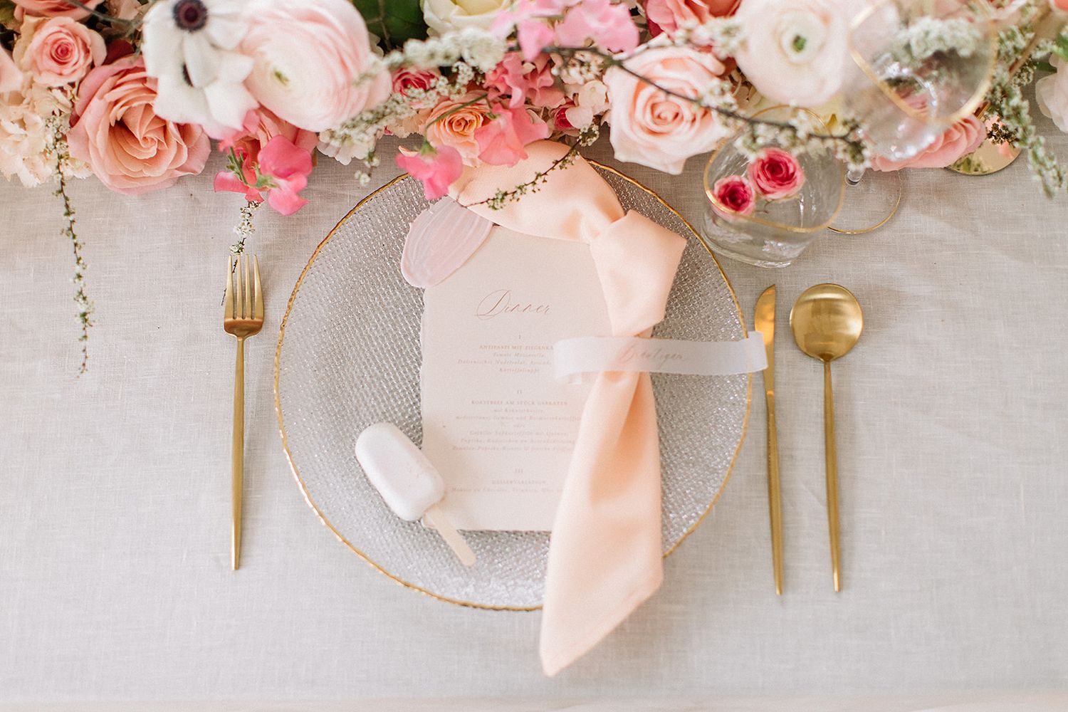 Elegante Platzdekoration mit goldenem Besteck, pfirsichfarbener Serviette und rosa Blumenzentrum auf beiger Tischdecke.