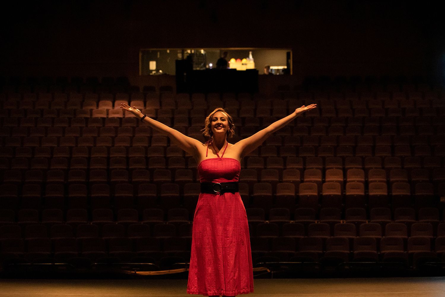 Photography of erformer in a red dress stands on stage with arms raised in an empty theater.