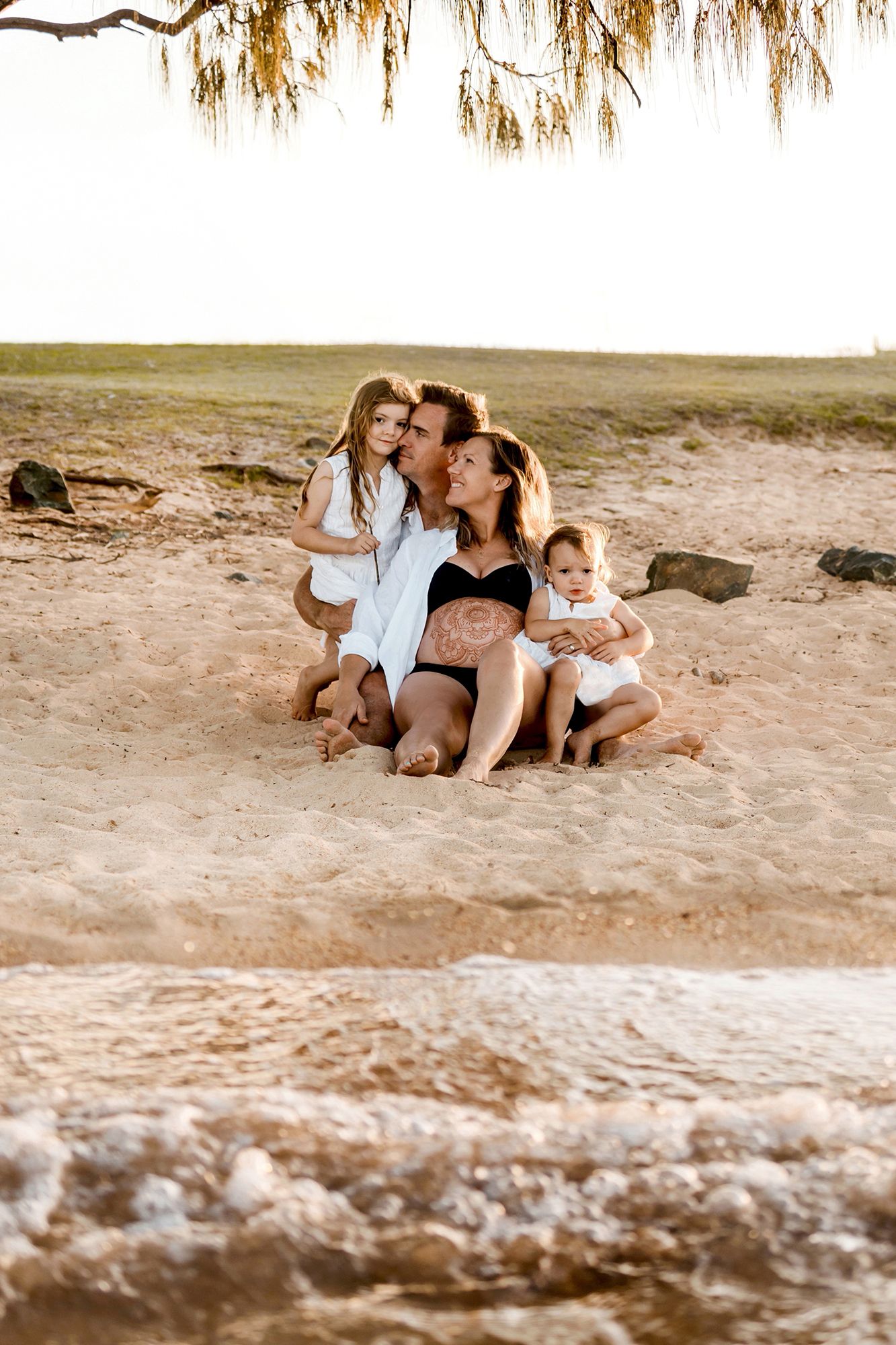 Family photography including a pregnant mother sit together on a sandy beach at sunset in Brisbane's bayside suburb of Raby Bay.