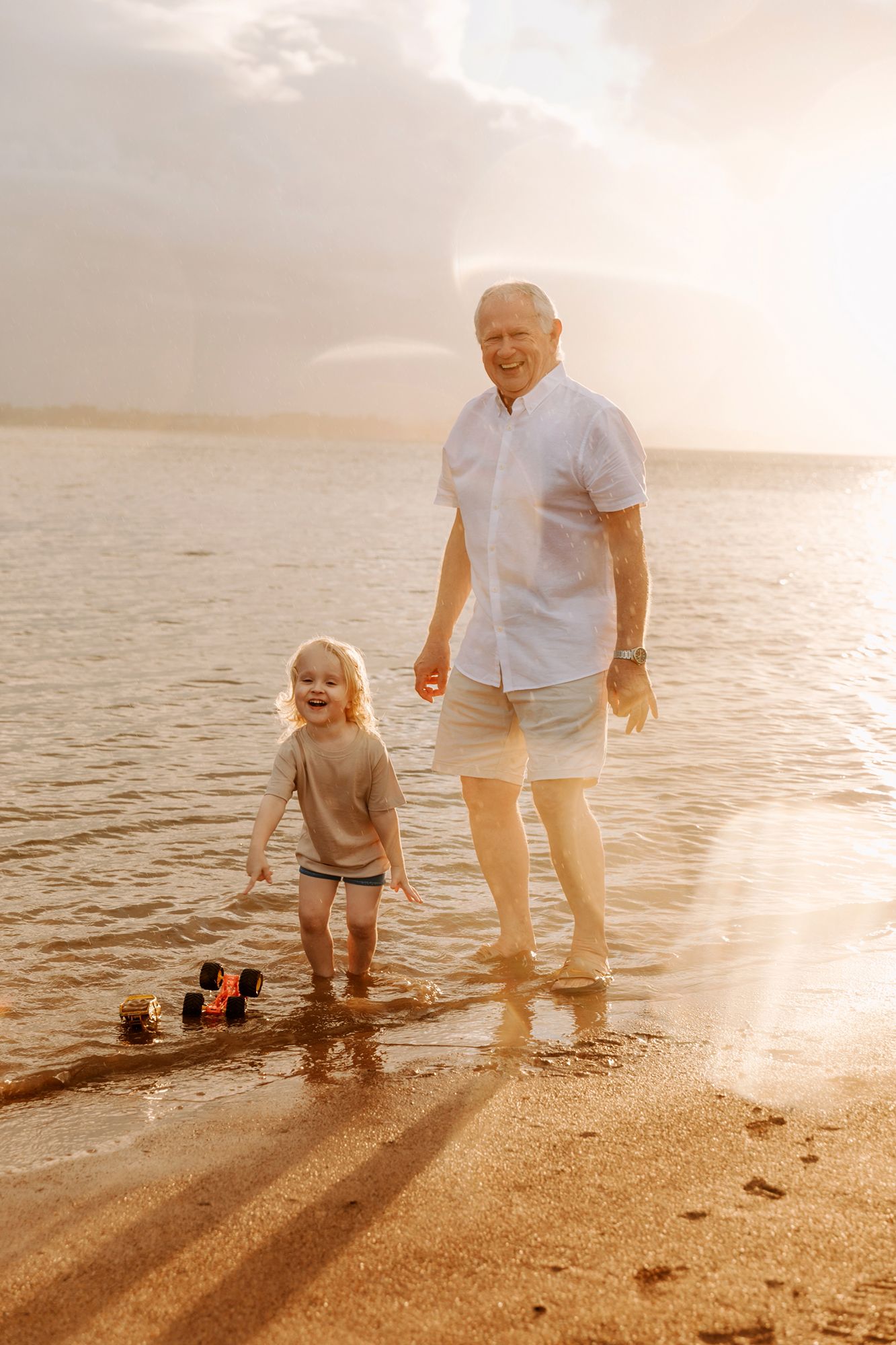 Grandfather and grandson walk along a sandy beach at sunset while playing in the gentle waves at Brisbane bayside beach of Wellington Point.