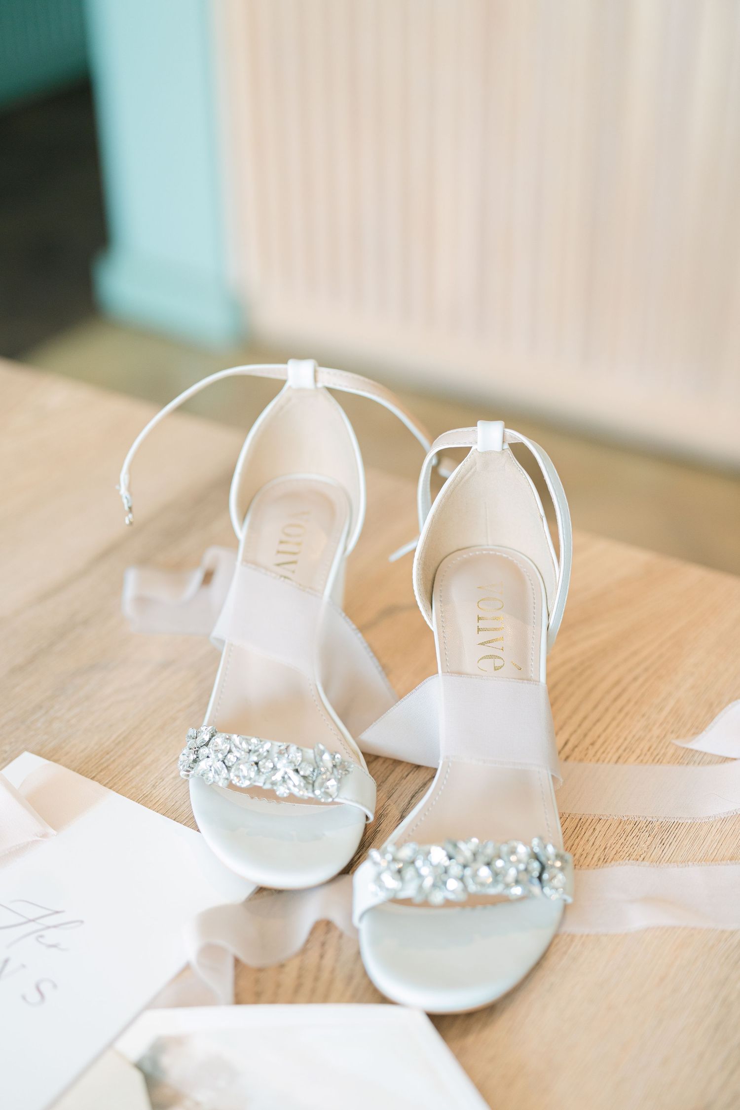 A sequence of white bridal shoes with crystal embellishments lined up on a wooden floor against a pale wall.